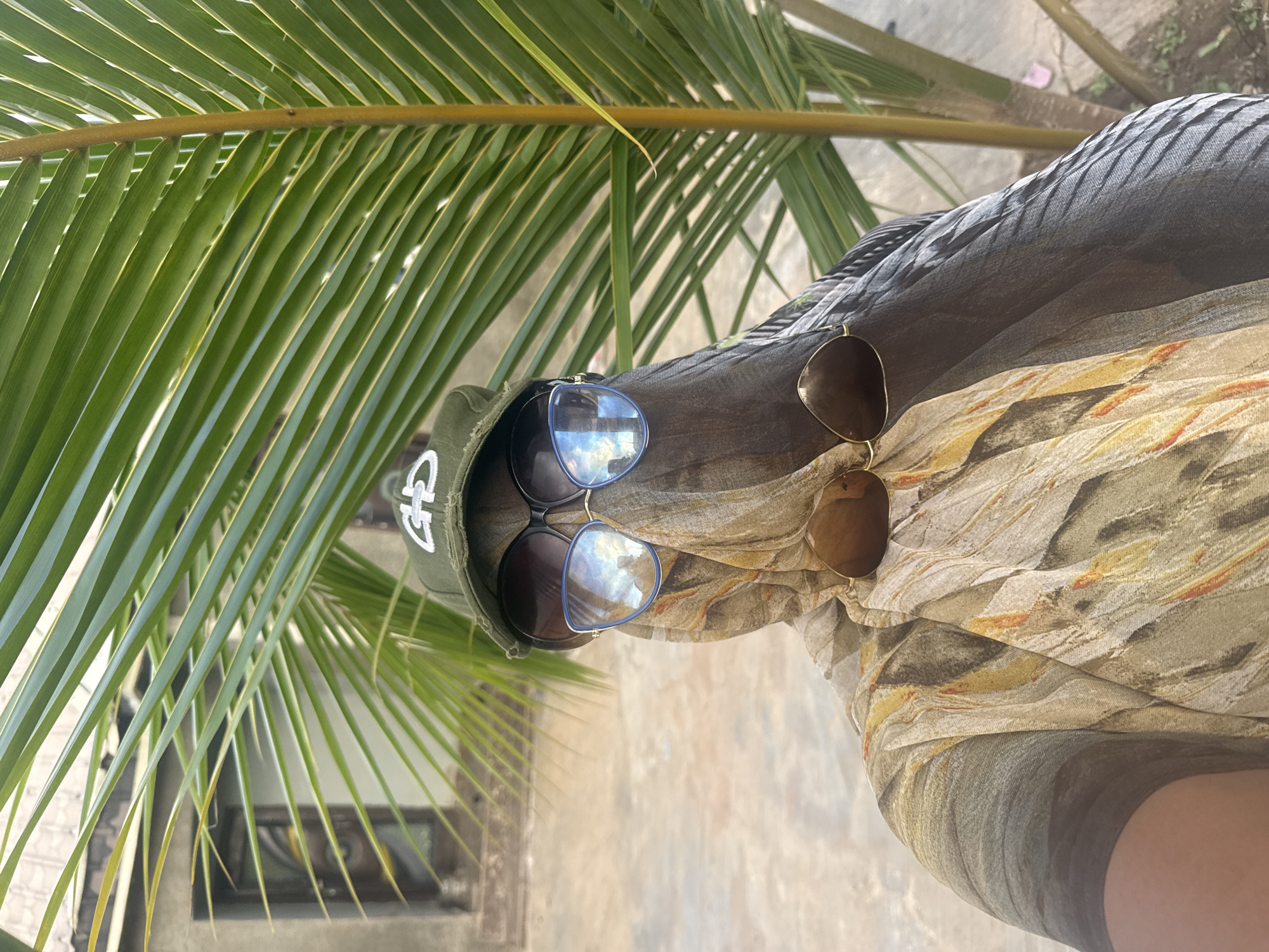In Jawu, Nigeria, a person is photographed outdoors, partially obscured by a large palm frond. The individual wears a green baseball cap with a white logo, large dark sunglasses, and a patterned fabric that covers their face and head, with another pair of brown sunglasses resting on the fabric. The background shows a building with windows and a light-colored, textured ground.