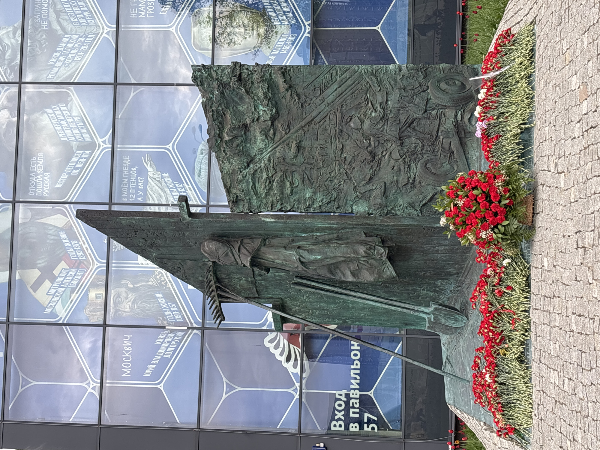 The image depicts a bronze monument in Moscow, Russia. The monument features a depiction of a peasant woman with a rake. Behind the monument is a wall with text in Cyrillic, including "Moscow," and historical information. The base of the monument is adorned with red and white flowers. A sign near the base of the monument reads "Entrance to pavilion 57." The scene is partially situated on a paved area.