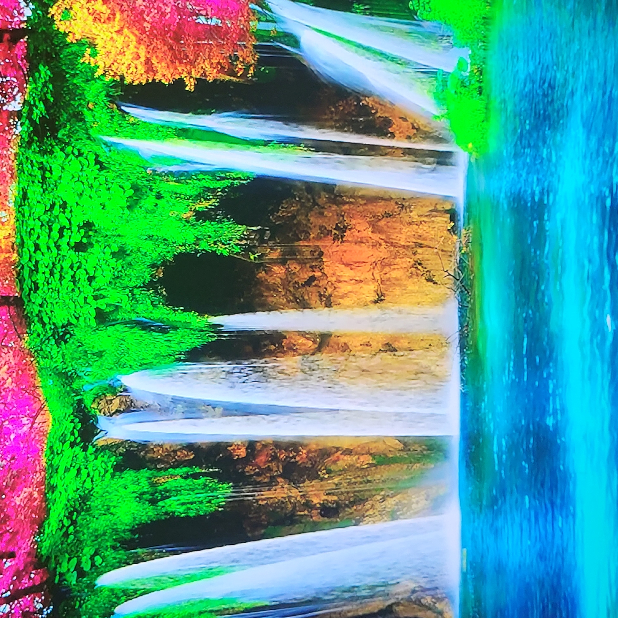 The image shows a vibrant, abstract representation of a waterfall cascading into a pool of water. The surrounding landscape is depicted with intense, unnatural colors: pink and yellow foliage on the left, transitioning to green, with amber and brown areas mid-frame, and a turquoise-blue body of water to the right. The waterfall itself is a series of multiple cascades, depicted as streaks of white with blue hues. The scene appears to be inverted. It is unlikely that this image accurately represents a real-world location, and its abstract nature makes a precise identification of a specific place impossible. If pressed to assume a location of the city of Yerevan, Armenia, a possible candidate could be an image or artistic interpretation of the waterfall in the Yerevan Botanical Garden, but with artificially manipulated colors.