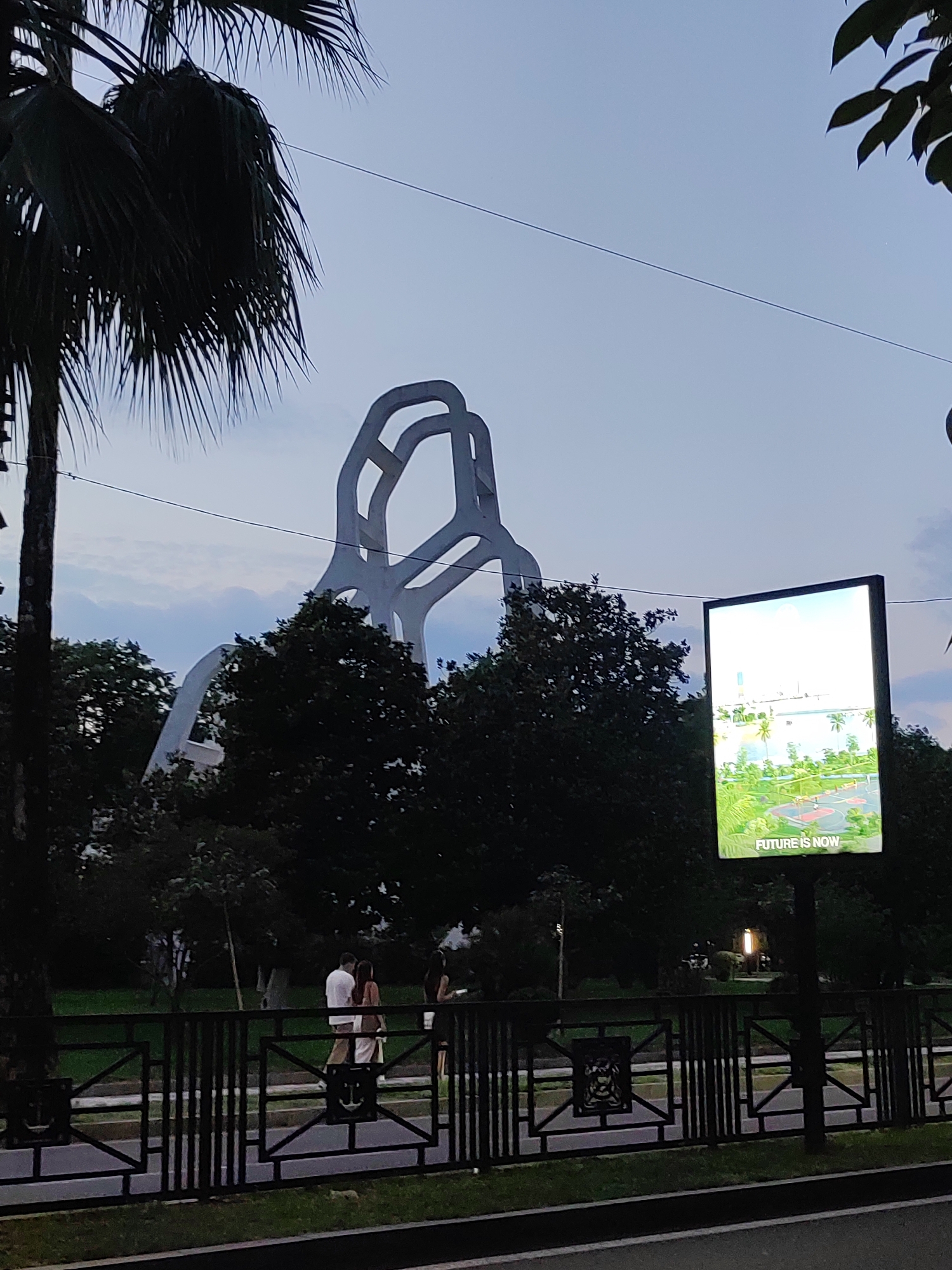 This image captures a street scene in Batumi, Georgia, at dusk. In the background, a large, abstract white sculpture dominates the skyline, with its looping, interconnected forms silhouetted against the twilight sky. In the middle ground, dark green trees partially obscure the sculpture. To the right, a brightly lit digital billboard displays a vibrant image of a modern city development, with the text "FUTURE IS NOW" visible at the bottom. In the foreground, a black metal fence runs horizontally across the scene. Behind the fence, several people are walking, including a couple in the center wearing light-colored clothing, and a woman in dark clothing to the right. A palm tree is prominent on the left side of the frame. The overall atmosphere is serene, with the fading light creating a soft, diffused illumination.