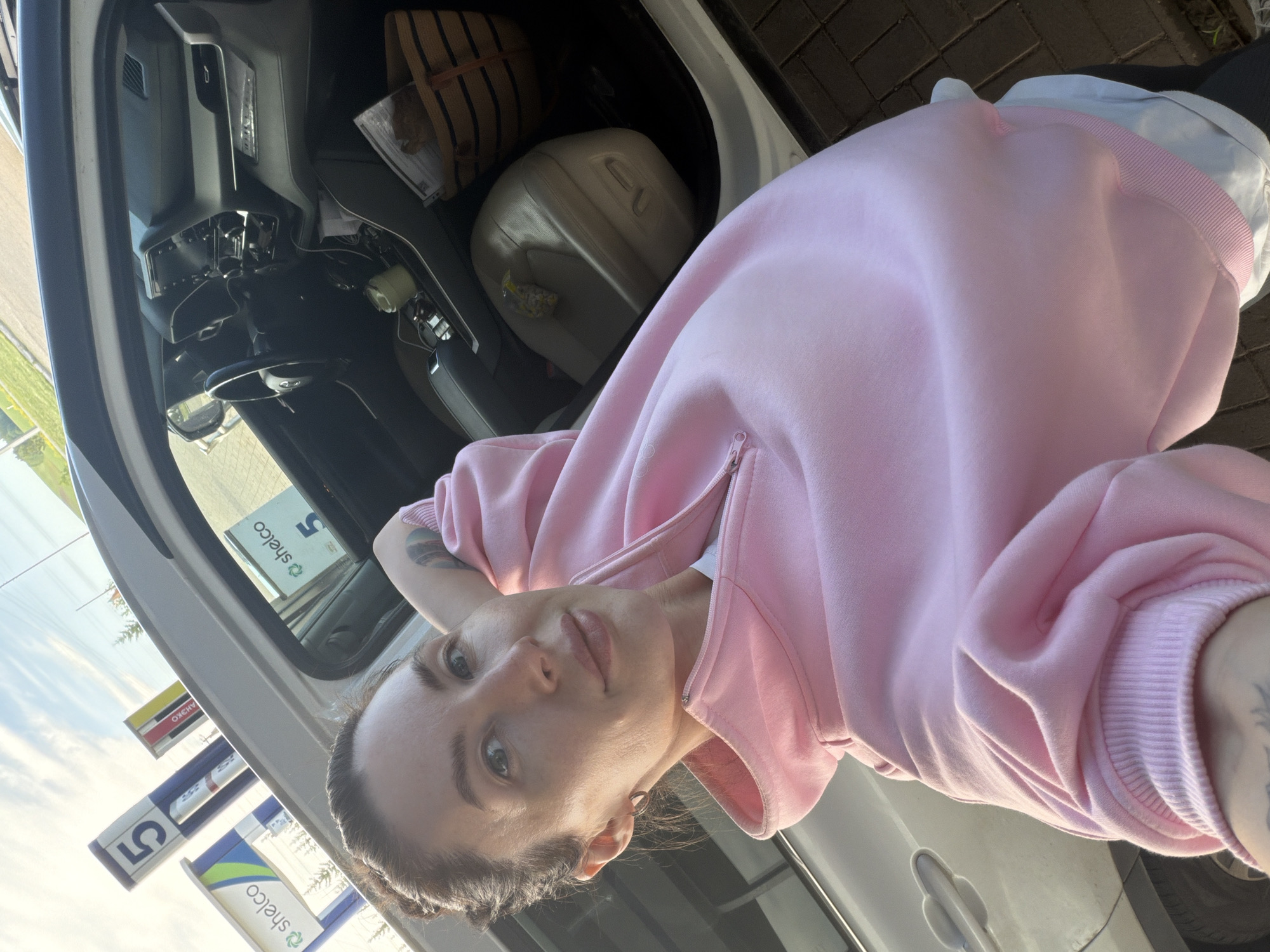 The image is a selfie taken by a woman inside a car, likely at a gas station in Ильинское, Russia. The woman, who appears to be young to middle-aged, is positioned diagonally across the frame, looking directly at the camera with a neutral to slightly smirking expression. She has light-colored eyes and her hair is styled in braids pulled back. She is wearing a pink full-zip hooded sweatshirt over a white undershirt, and a tattoo is visible on her left arm. Small hoop earrings adorn her ears.
The car's interior is partially visible, showing the dashboard, steering wheel, and part of the front passenger area. On the passenger seat or in the rear, a striped straw-like tote bag, a light-colored jerrycan or large plastic container, and a yellow wrapper are present.
Outside the car window, several gas station signs are visible under a bright, clear daytime sky. Prominently displayed are signs featuring the brand name "shelco" in green and blue logos, along with the number "5". Another yellow sign shows the text "АНЭКО" (ANEXO). The ground outside is paved with bricks. The scene suggests a casual stop, possibly for refueling, during clear weather.