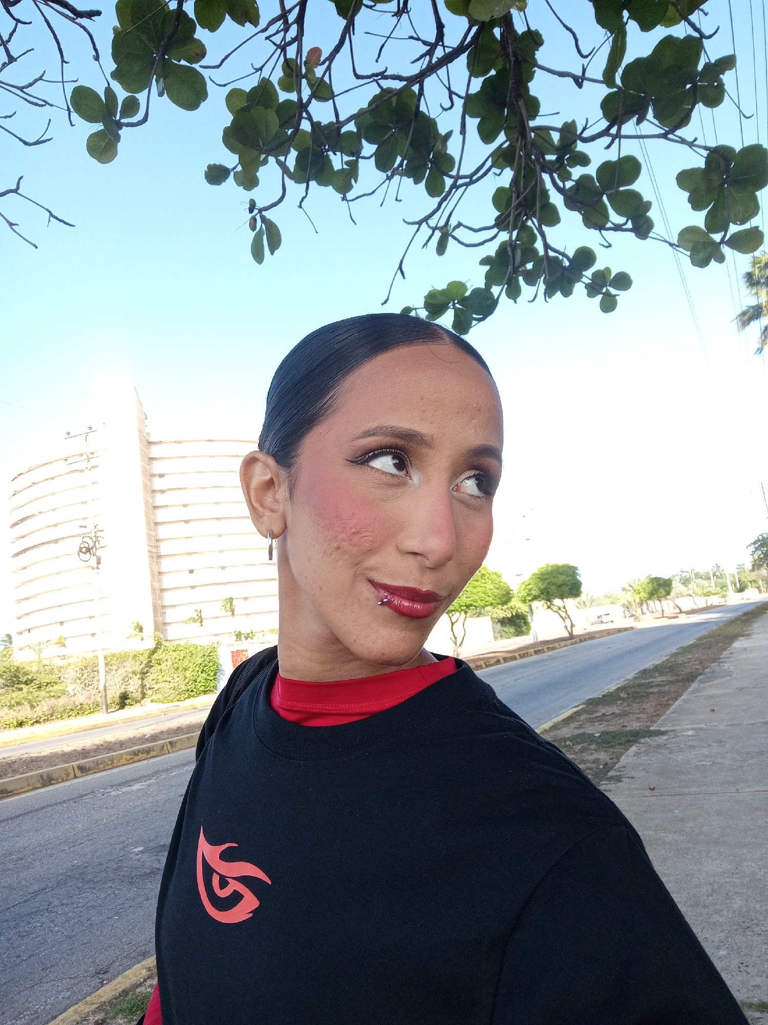 A medium shot depicts a woman standing near a street in Porlamar, Venezuela. She is fair-skinned with dark hair pulled back, wearing a black t-shirt with a red logo, and a red undershirt. She has a lip piercing and is looking off to the side. Above her, there are tree branches with green leaves against a blue sky. To the left, there is a large white building and a fence covered in greenery. A road stretches out in front of her, with a sidewalk on the right side of the frame.