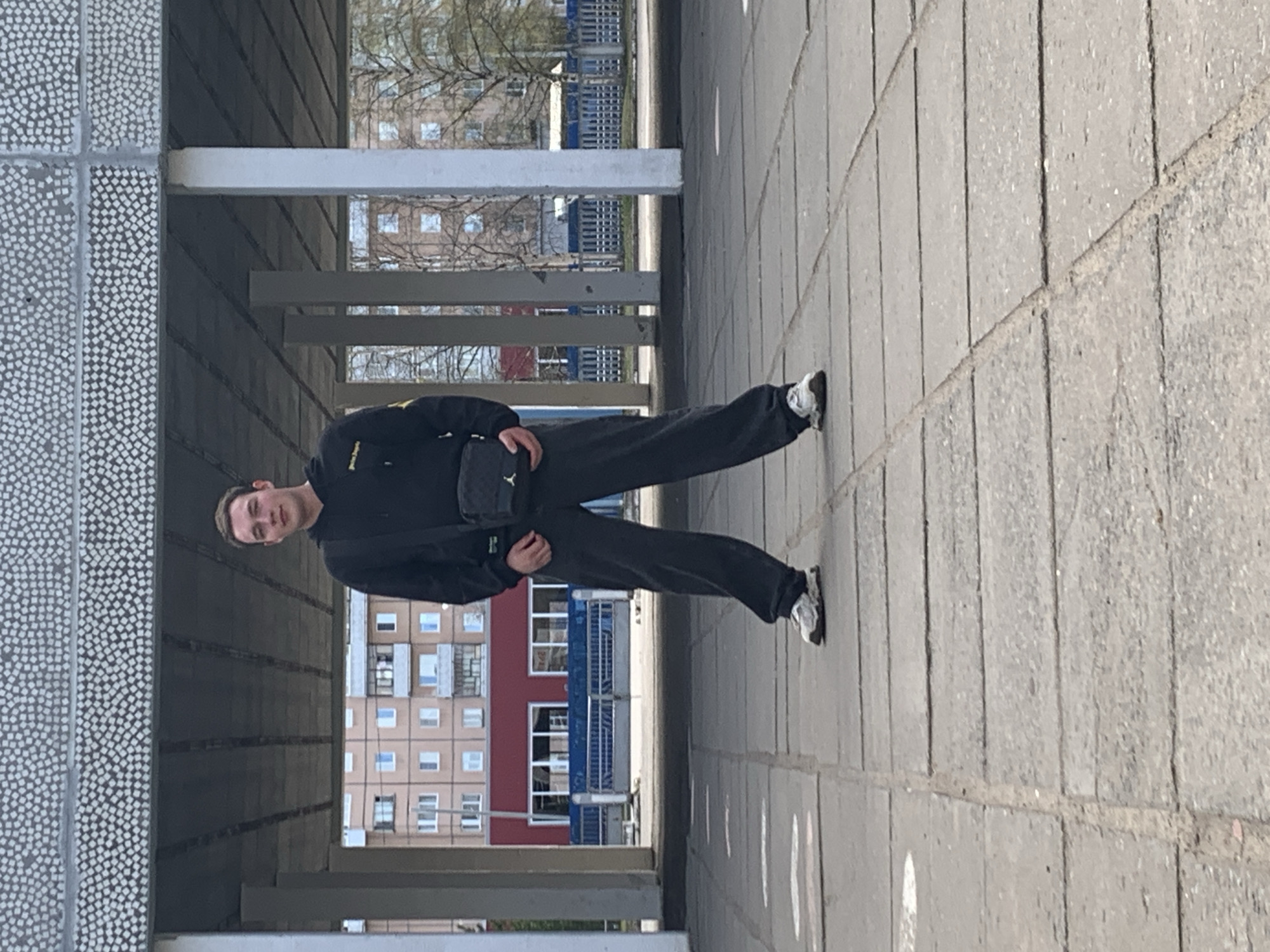 A young man stands under a pedestrian overpass in Severodvinsk, Russia. He is wearing a black jacket, dark pants, and light-colored sneakers. He has a small black shoulder bag. The overpass has a textured, patterned wall on one side, with a series of columns and railings. In the background, multi-story apartment buildings are visible. The ground is paved with concrete blocks.