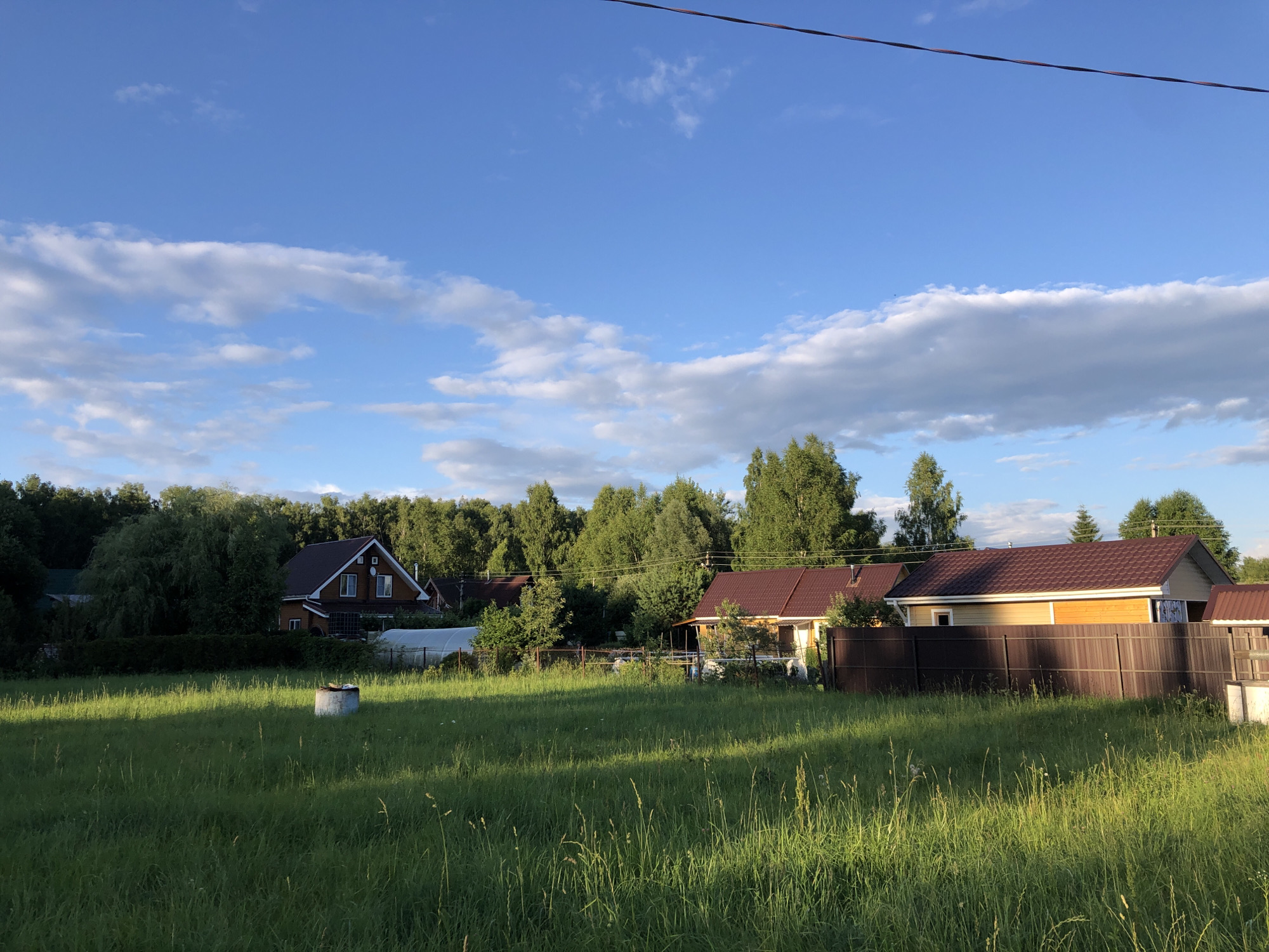 The image depicts a view of residential buildings and a grassy field under a bright blue sky with scattered white clouds. The setting appears to be the city of Московский Ветеран Снт, Russia, characterized by lush green vegetation and several houses with dark-tiled roofs. In the foreground, a large, uncultivated grassy area dominates the scene, with tall blades of grass catching the sunlight, creating dappled patterns. A circular, concrete structure, possibly a wellhead or a decorative element, is visible in the middle of the field.
Behind the field, a row of houses is situated, bordered by a dark brown fence. One house, painted in a bright yellow hue with a dark tiled roof, is prominently featured. To its left, another house with a reddish-brown roof and wooden siding is visible, set against a backdrop of dense green trees. A greenhouse with a white translucent cover is located between these houses. The background is filled with a thick line of mature trees, suggesting a semi-rural or suburban environment. A power line stretches across the upper part of the sky. No people are visible in the image. The overall impression is of a peaceful, sun-drenched summer day in a residential area surrounded by nature.