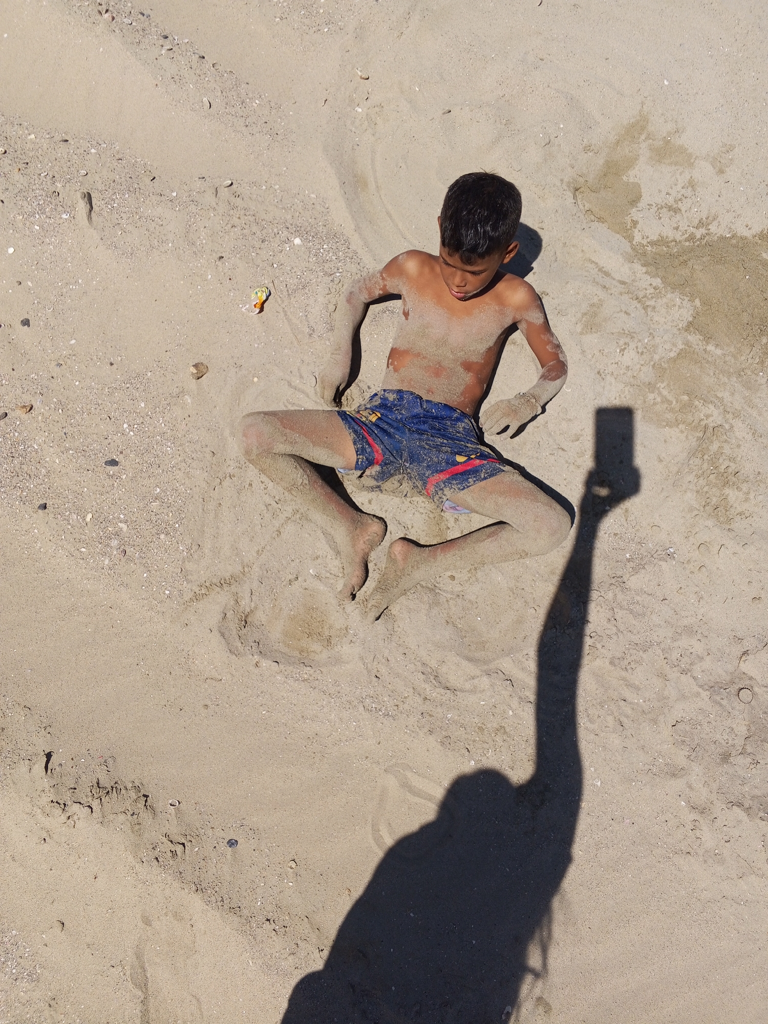 A high-angle shot depicts a young boy lying on a sandy beach. He is wearing blue swim trunks with red and pink accents. His body and the trunks are covered in sand. The shadow of a person holding a phone stretches across the lower right portion of the image. The sand is a light brown color, and scattered debris, including a small piece of orange trash, is visible. The scene is sunlit, suggesting a bright, clear day. This image was likely captured near the city of Juan Griego, Venezuela.