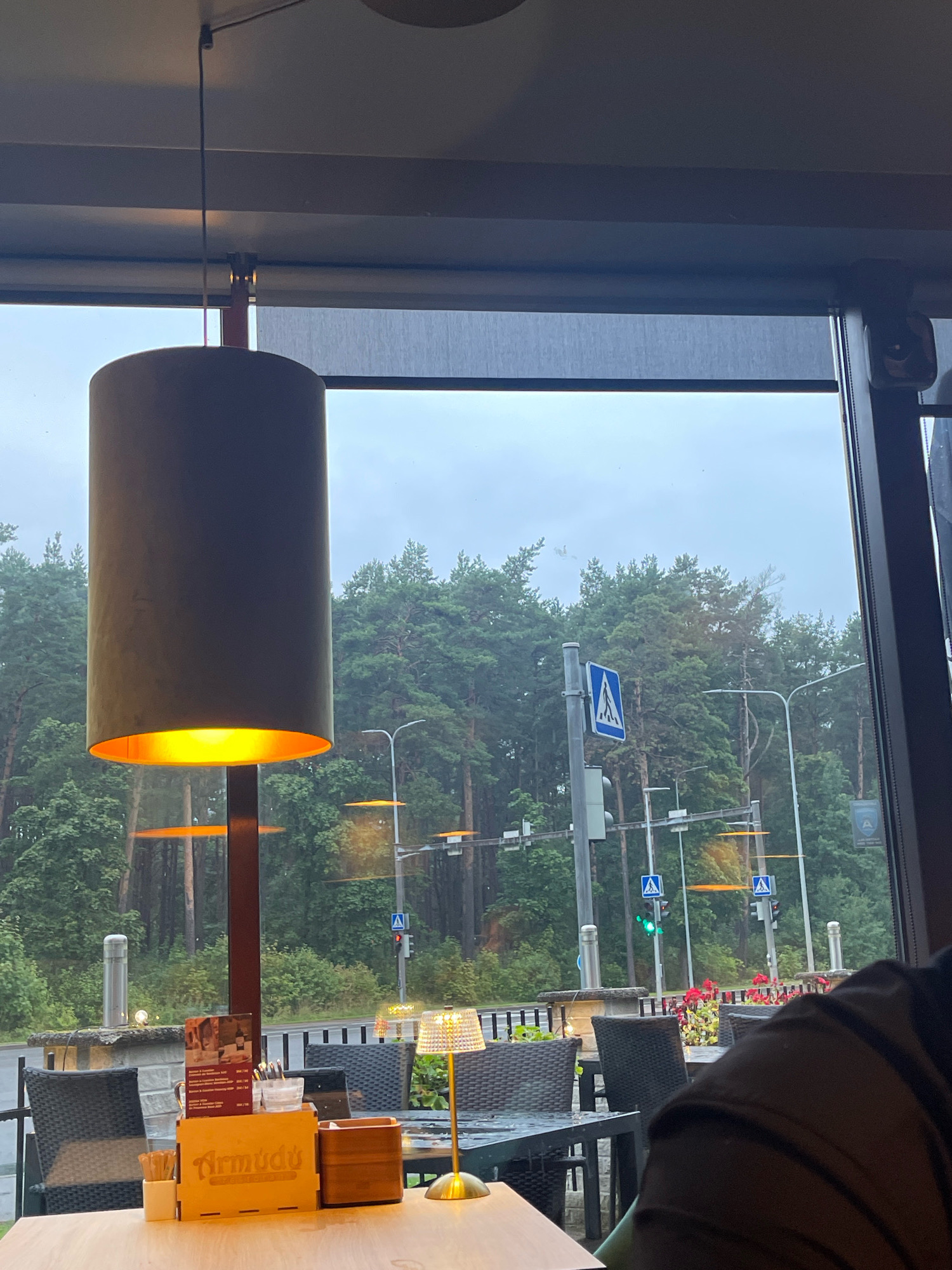 The image is a close-up view from the inside of a cafe or restaurant in Tallinn, Estonia, looking out through a large window. In the foreground, a large, cylindrical pendant lamp with a warm yellow light is hanging from the ceiling, partially obscuring the view. A wooden table in front of the window holds a menu, cutlery, and a small table lamp. Outside, a road with traffic lights and trees is visible. One traffic light shows a green signal, indicating that traffic can proceed. A pedestrian crossing sign is also visible. The overall scene suggests a rainy or overcast day.