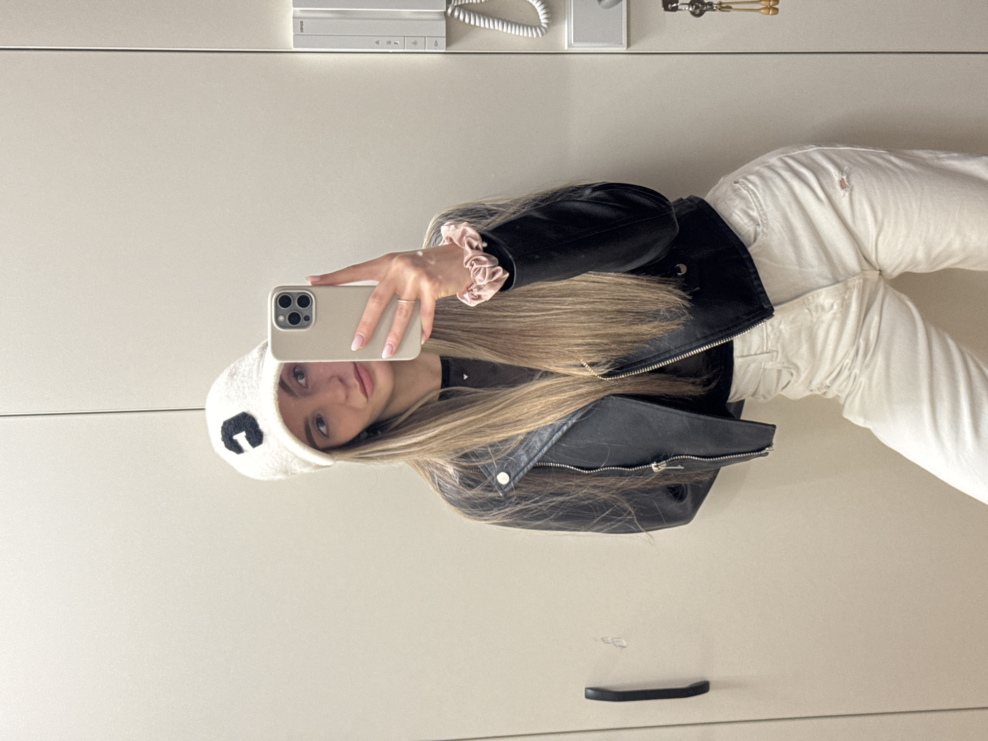 A young female individual, estimated to be in her late teens or early twenties, is depicted in a full-length mirror selfie. She has long, straight, light brown hair with blonde highlights. She is wearing a white textured baseball cap featuring a black capital 'C' emblem on the front, a black faux leather motorcycle jacket with prominent silver zippers and snaps, a dark (possibly black or sheer) top underneath, and high-waisted off-white denim jeans with a small tear on the upper left thigh. On her right wrist, she wears a pale pink satin scrunchie, and a gold-toned ring is visible on the fourth digit of her right hand. She is holding a light gold-colored iPhone, identifiable by its triple rear camera array, to capture the image. The background consists of light beige wall panels or built-in cabinetry. On the left, a black horizontal handle suggests a door or drawer, accompanied by a faint scuff mark. On the right, a white wall-mounted intercom system with multiple buttons and a coiled cord leading to a handset is present, below which is a white rectangular utility panel. Further to the right, a set of keys adorned with two ornate gold-colored keychains hangs from a hook. The environment is consistent with a modern residential or commercial interior in an urban setting, likely in Sofia, Bulgaria.