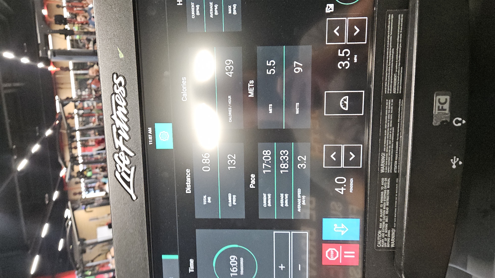 The image provides a close-up view of a Life Fitness treadmill's digital console, displaying workout statistics within a gym environment in Plovdiv, Bulgaria. The time shown on the screen is 11:07 AM.
The console indicates an ongoing workout session with a recorded time of 16 minutes and 9 seconds. The user has covered a total distance of 0.86 miles and climbed 132 feet. The current pace is 17 minutes and 8 seconds per mile, while the average pace is 18 minutes and 33 seconds per mile, corresponding to an average speed of 3.2 miles per hour. Other metrics include an estimated 439 calories burned per hour, 5.5 METs, and 97 Watts. The treadmill is currently set to a speed of 3.5 MPH and an incline (PENDENZA) of 4.0.
Below the main display, control buttons for "STOP" and "PAUSE" are visible, along with directional arrows for adjusting speed and incline. The "Life Fitness" logo is prominently displayed on the console's frame. The background is blurred but shows rows of other gym equipment under bright artificial lighting, typical of a fitness center.
Several safety warnings are present on the right side of the console in both English and French. These texts advise users to read the instruction manual, exercise extreme caution when stepping onto or off a moving belt, inspect equipment before use, and keep children under 13 away from the machine to prevent injury. Icons for USB and headphone connectivity are also visible.