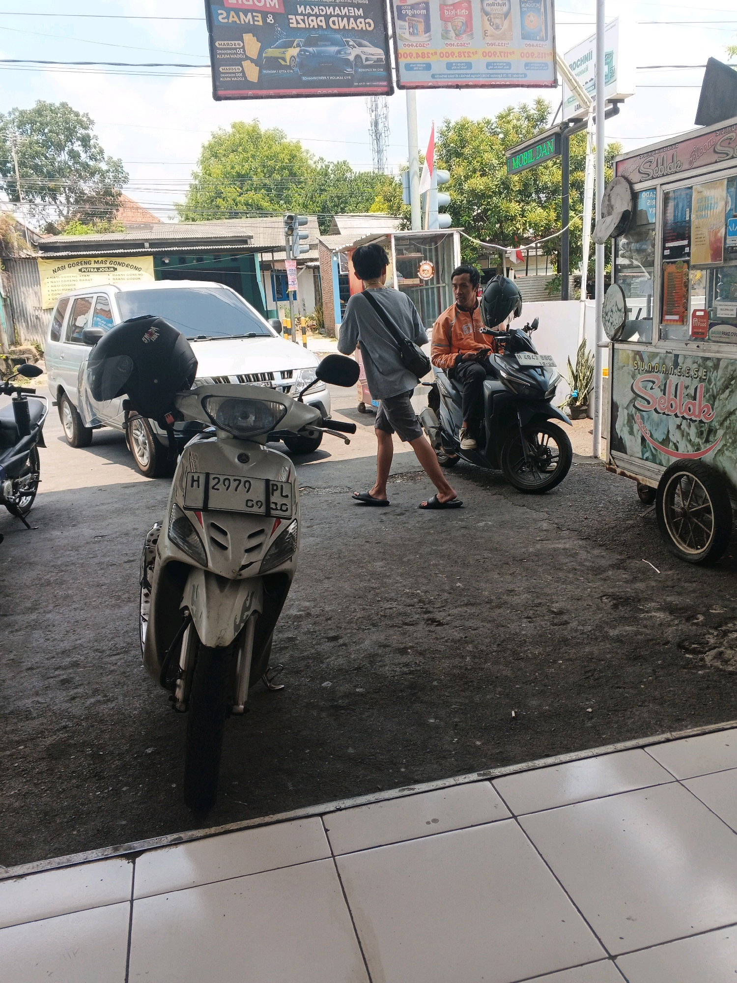 In the city of Genuk Barat, Indonesia, a street scene unfolds with several people and vehicles. A white scooter with license plate "H 2979 PL" is parked prominently in the foreground, a black helmet resting on its handlebars. Behind it, a silver SUV is partially visible. To the right, a man in an orange jacket sits on a black motorcycle, facing forward, while another person in a grey shirt and shorts walks away from the viewer towards the left. A food cart, with the name "Sedlak" prominently displayed, is situated to the right of the motorcycles. The environment appears to be a street or parking area with a rough asphalt surface and some paved tiles in the foreground. Overhead, signage indicates commercial activity. The sky is bright, suggesting daylight.