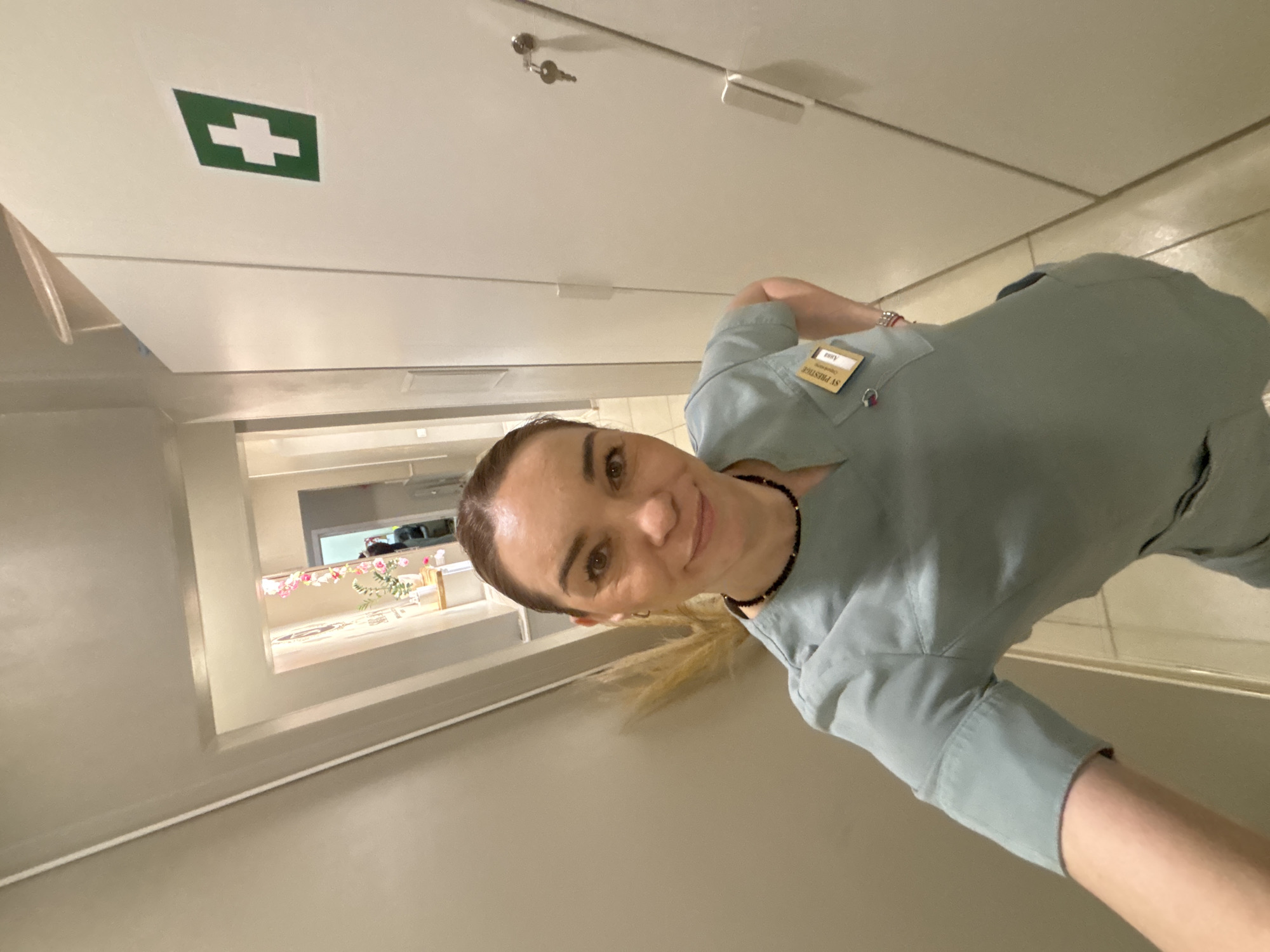 A woman is taking a selfie in a hallway. A green medical cross is visible on the ceiling, suggesting the location is a healthcare facility. The woman is wearing a light blue uniform with a name tag that reads "SV PRESTIGE." A hallway stretches into the background. In the background, a window or opening reveals a room with flowers and furniture. A set of keys hangs from the ceiling. The city of Vsevolozhsk, Russia, is not discernible from the image.