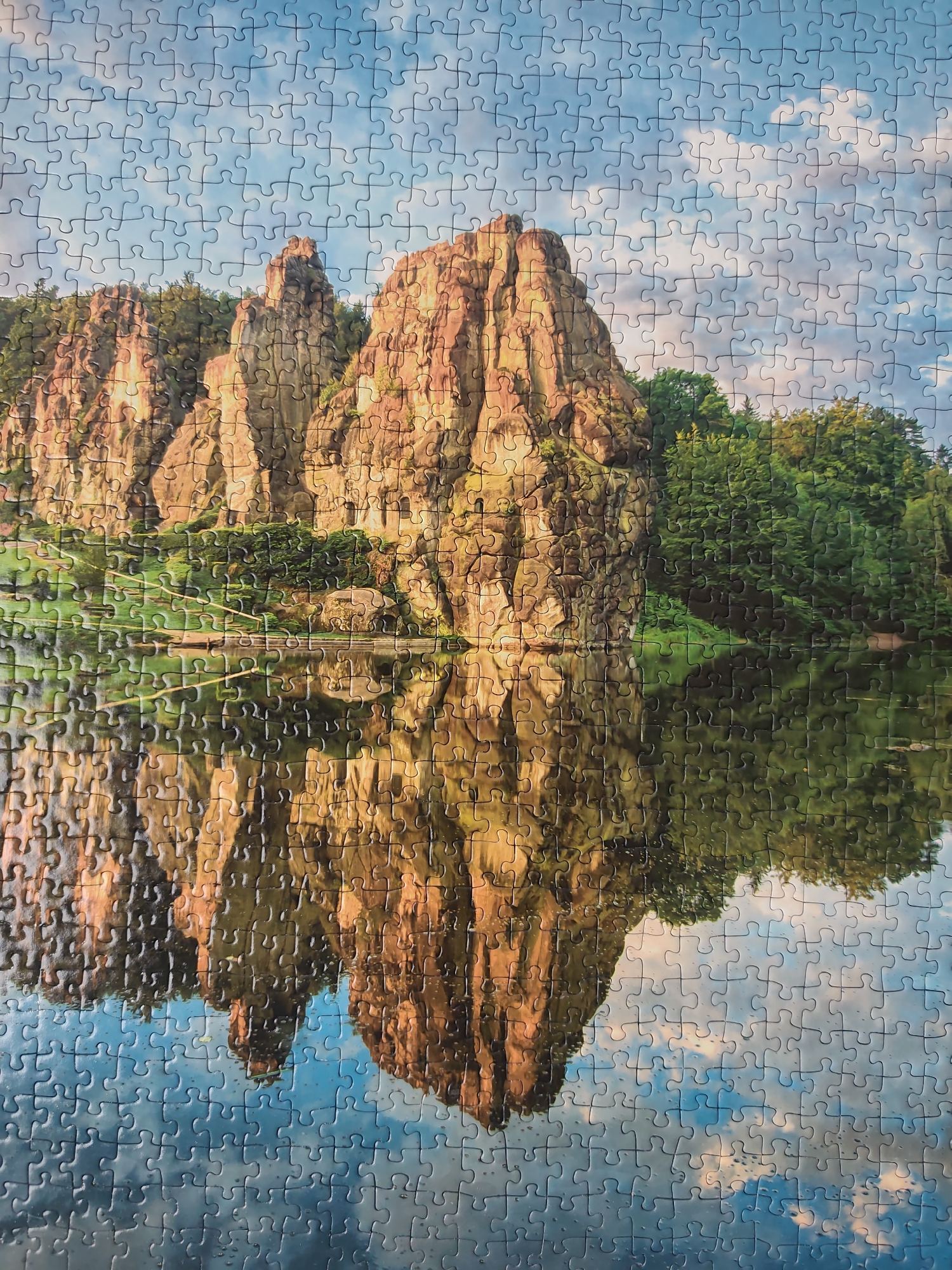 This is a jigsaw puzzle depicting the Externsteine, a distinctive rock formation in the Teutoburg Forest, near Horn-Bad Meinberg in North Rhine-Westphalia, Germany. The image shows the towering sandstone rocks reflecting in the calm water below. The sky is partially cloudy. Trees and greenery cover areas surrounding the rocks.