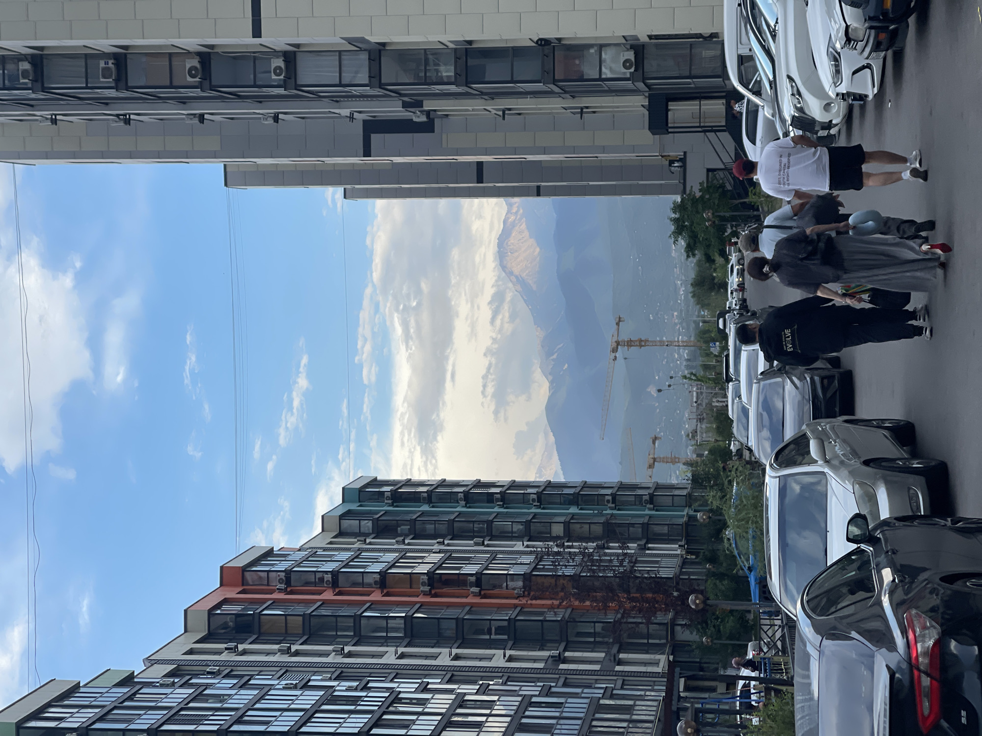 A group of people walks down a street in a modern residential district in Almaty, Kazakhstan, with a view of the Tian Shan mountains in the background. The street is lined with tall, multi-story apartment buildings. The building on the left has colorful accents of orange and teal, while the building on the right is a neutral grey and white. Several cars are parked along the street. Two construction cranes are visible in the distance, suggesting ongoing development. The sky is partly cloudy. One of the people in the group is carrying a blue inflatable toy. The overall scene depicts an urban environment with a natural landscape as a backdrop.