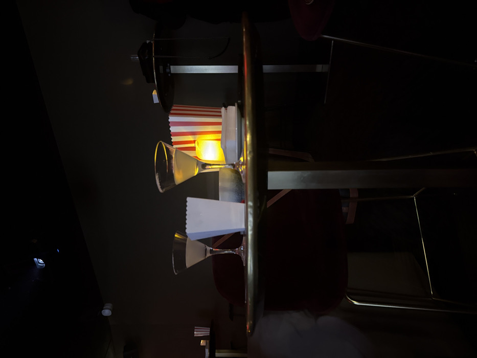 Two martini glasses and two popcorn containers are set on a golden bar cart. One popcorn container is white, the other has red and white stripes and is illuminated from behind. The cart has two shelves visible, and red seats are in the background. The scene is dimly lit.