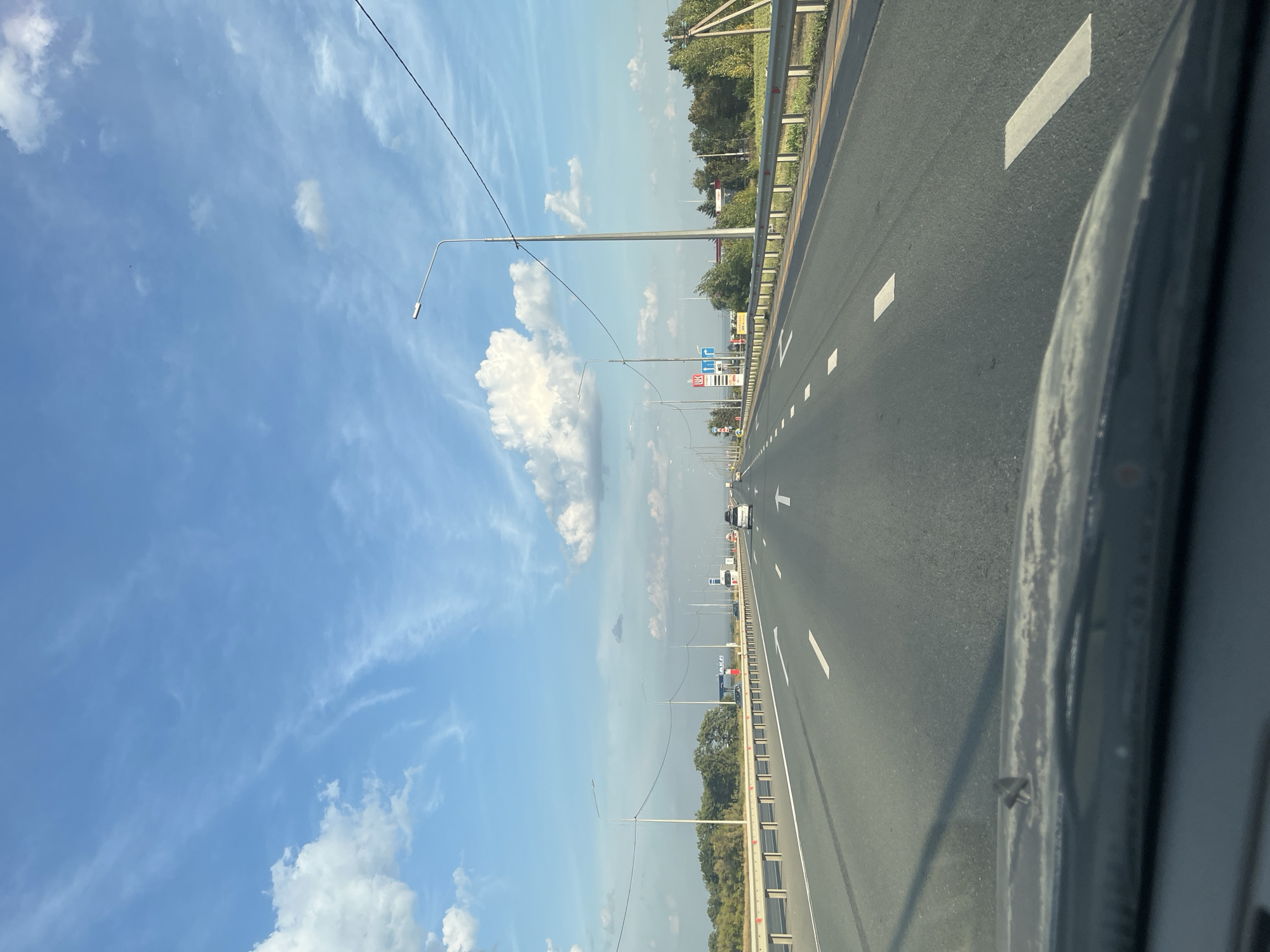 The image depicts a view from a vehicle traveling on a multi-lane highway in Dolgii Buerak, Russia. The highway stretches into the distance, lined with metal guardrails and streetlights. White lane markings are visible on the asphalt. The sky above is blue with scattered white clouds. Trees and vegetation form a treeline along the sides of the highway, with some structures, possibly related to a gas station, visible in the distance. A few other vehicles are present on the road. The foreground shows a portion of the vehicle's dashboard.