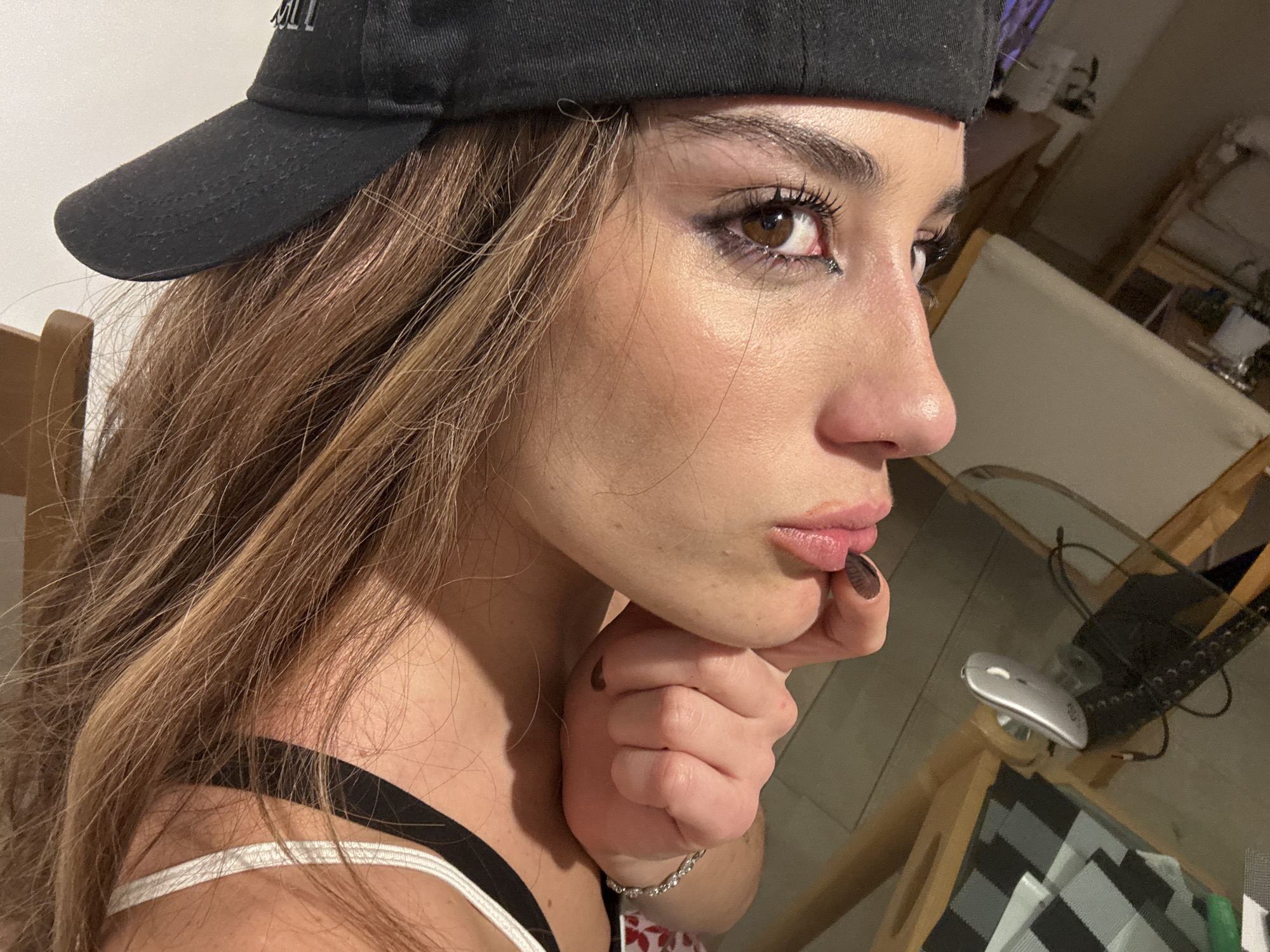 A young woman wearing a black baseball cap, white tank top with black straps, and a bracelet is resting her face on her fist. She has long brown hair and is wearing eye makeup. Her fingernails are painted black. The setting appears to be indoors, possibly a home. In the background is a chair, a table with a glass top, a computer mouse, and other items that are out of focus. There is no indication of the location being Buenos Aires, Argentina.