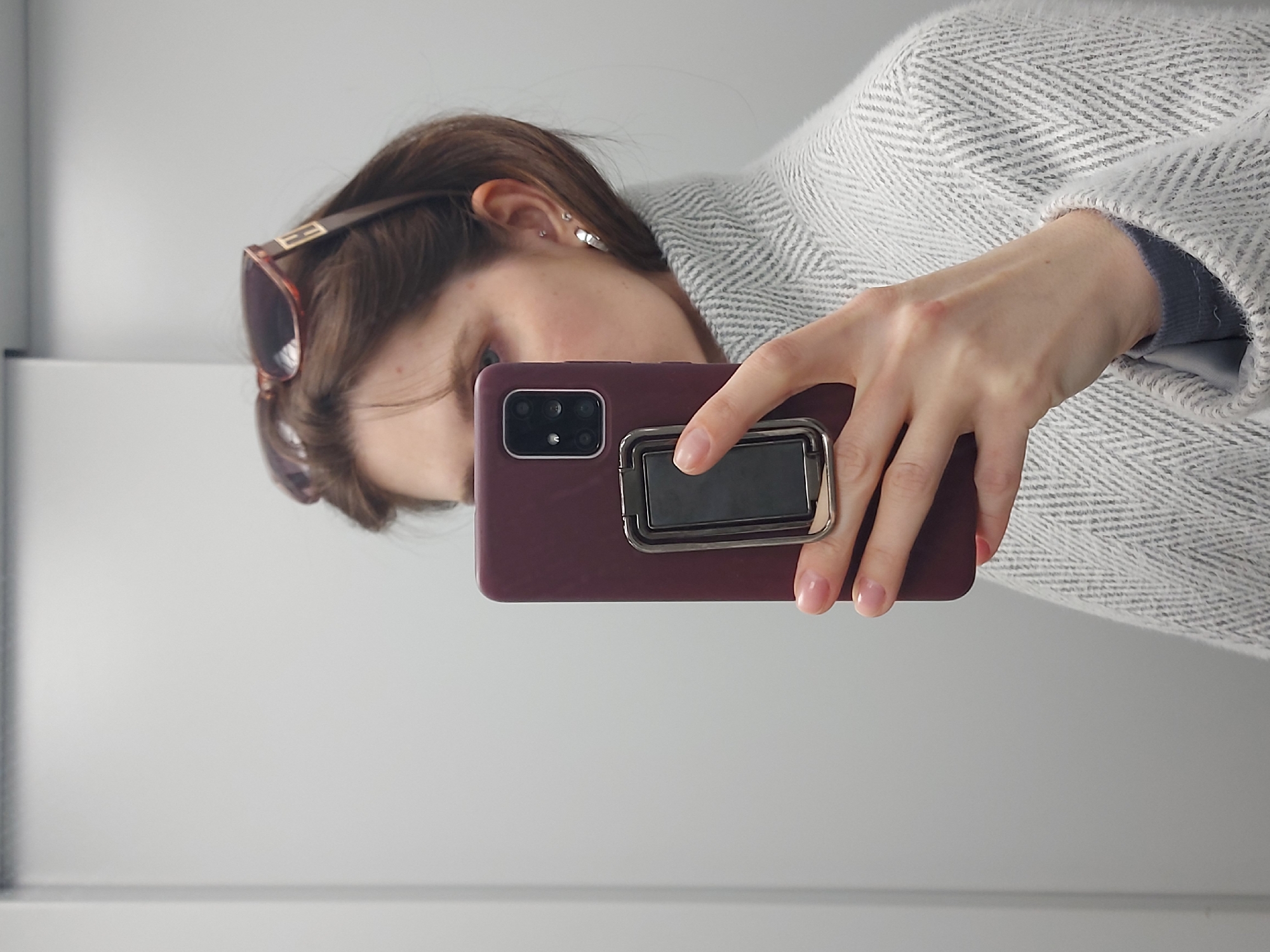 A person is taking a selfie in what appears to be a mirror. The person is wearing sunglasses and a textured, light-colored coat. They are holding a maroon-colored smartphone with a phone grip attached to the back. The background is a plain white surface. There is no information available in the image to indicate that this scene is located in the city of Chaikovsky, Russia.