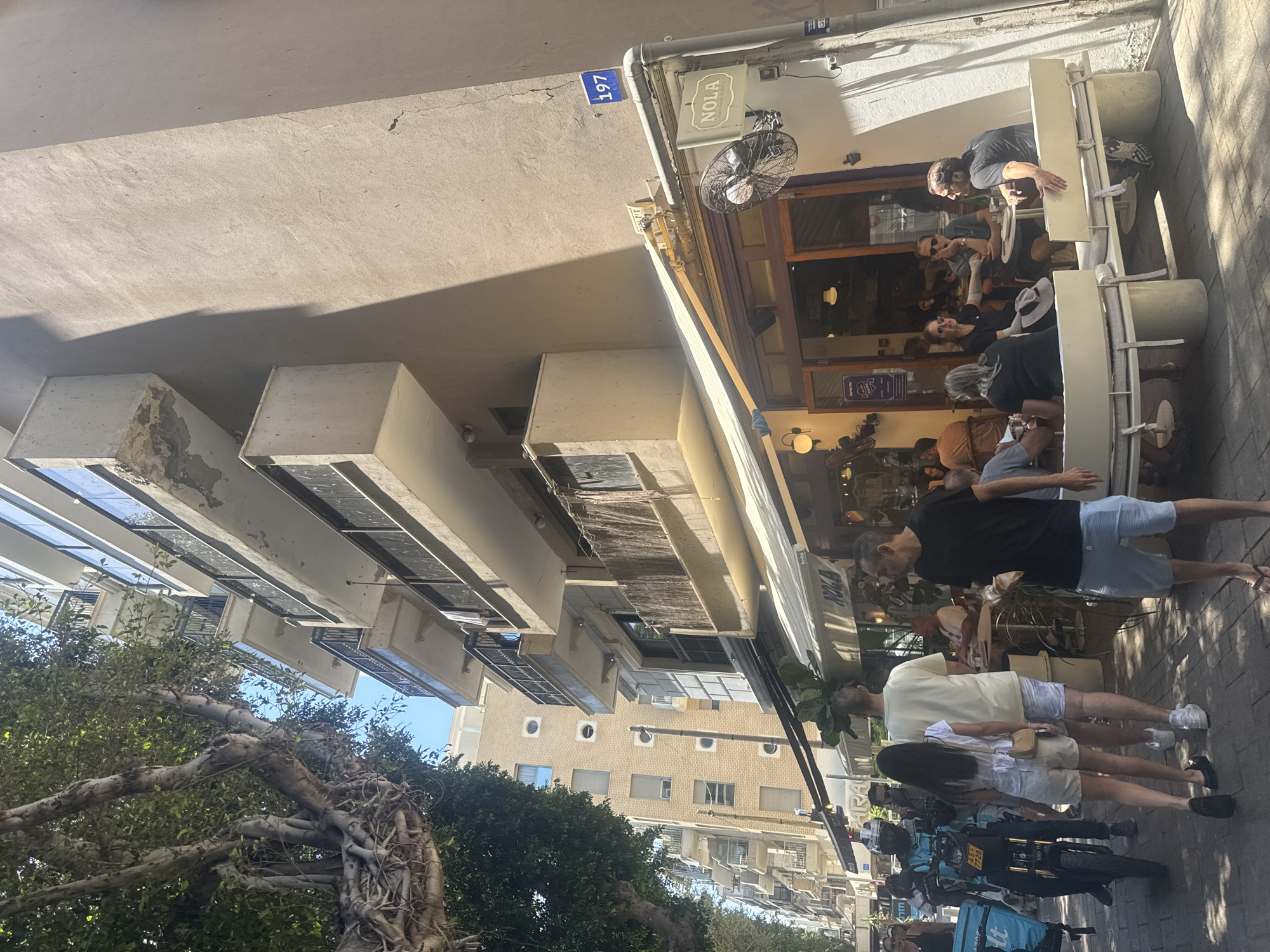The image shows a busy street scene outside a cafe in Tel Aviv, Israel. The cafe, named "NOLA," has outdoor seating occupied by patrons. A large number "197" is present on the building's exterior, likely indicating the street number. A black fan is mounted outside the restaurant.
Several people are seen walking along the sidewalk, including a couple and a delivery worker wearing turquoise clothing with a black delivery motor-scooter. Buildings of various heights are visible in the background, along with trees. The photograph captures a lively urban environment under bright daylight.