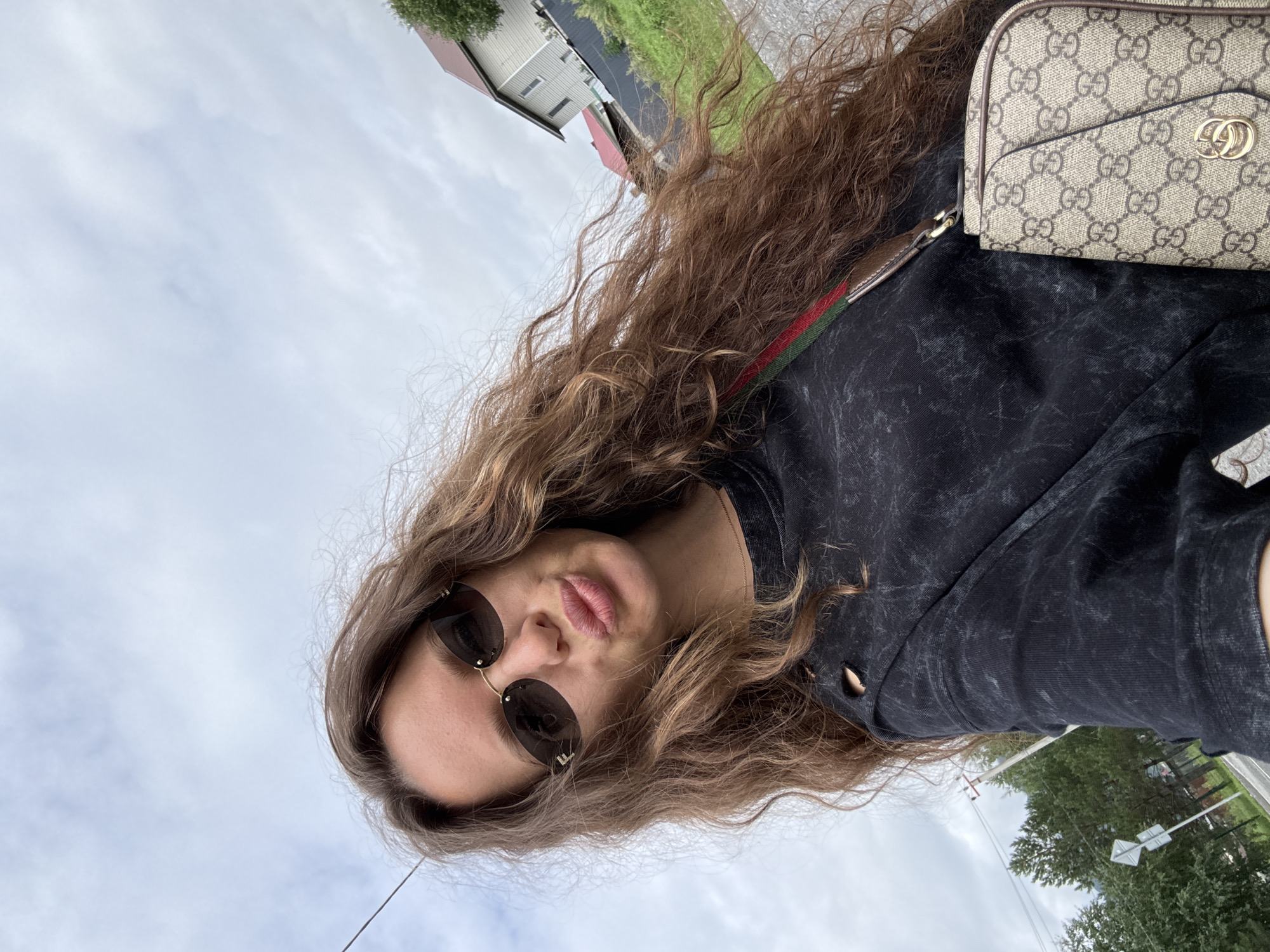 A young woman with long, wavy brown hair and sunglasses is taking a selfie outdoors. She is wearing a dark-colored, possibly denim, long-sleeved shirt with distressed detailing. A Gucci-patterned crossbody bag with red and green straps is visible. Her lips are pursed. The background shows a cloudy sky, some houses with red roofs, green vegetation, and trees, suggesting a rural or suburban setting. The presence of a power line in the upper left corner indicates some infrastructure. The scene is likely in or near Chemal, Russia, a region known for its natural beauty and tourist attractions.