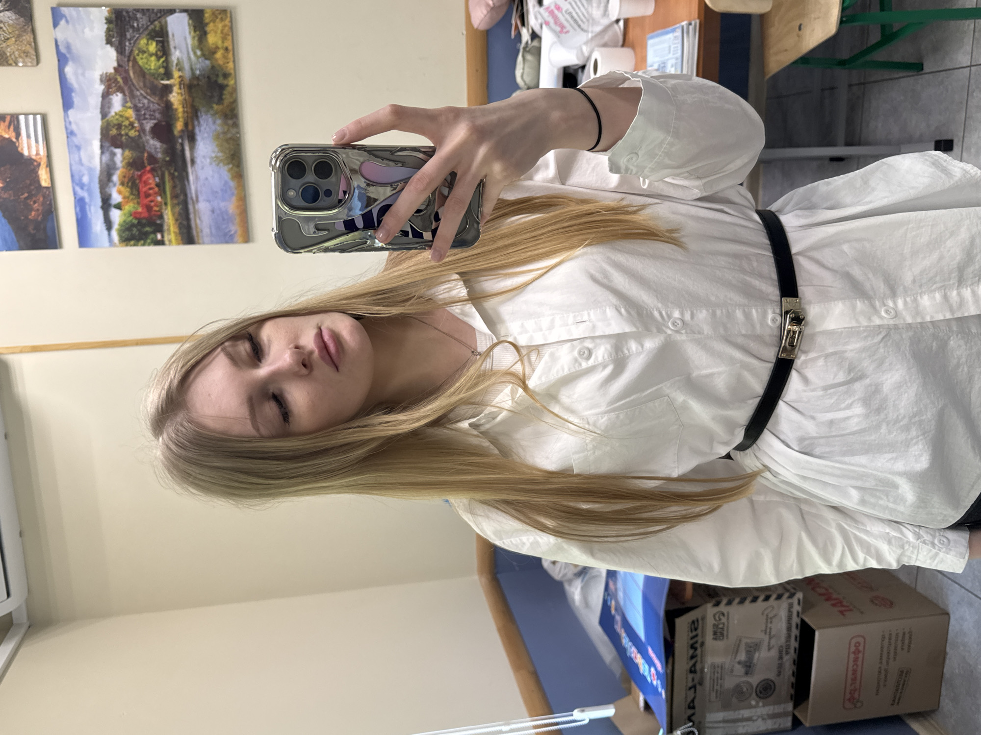 A fair-skinned, long blonde-haired woman is taking a selfie with a silver-colored smartphone, wearing a white collared button-down shirt with a thin black belt. The woman's eyes are closed. Two framed pictures hang on the wall in the background. A desk and chair are partially visible in the upper right corner. Brown cardboard boxes and blue and white objects are on the floor below. Based on the text on the boxes, the location is Perm, Russia.