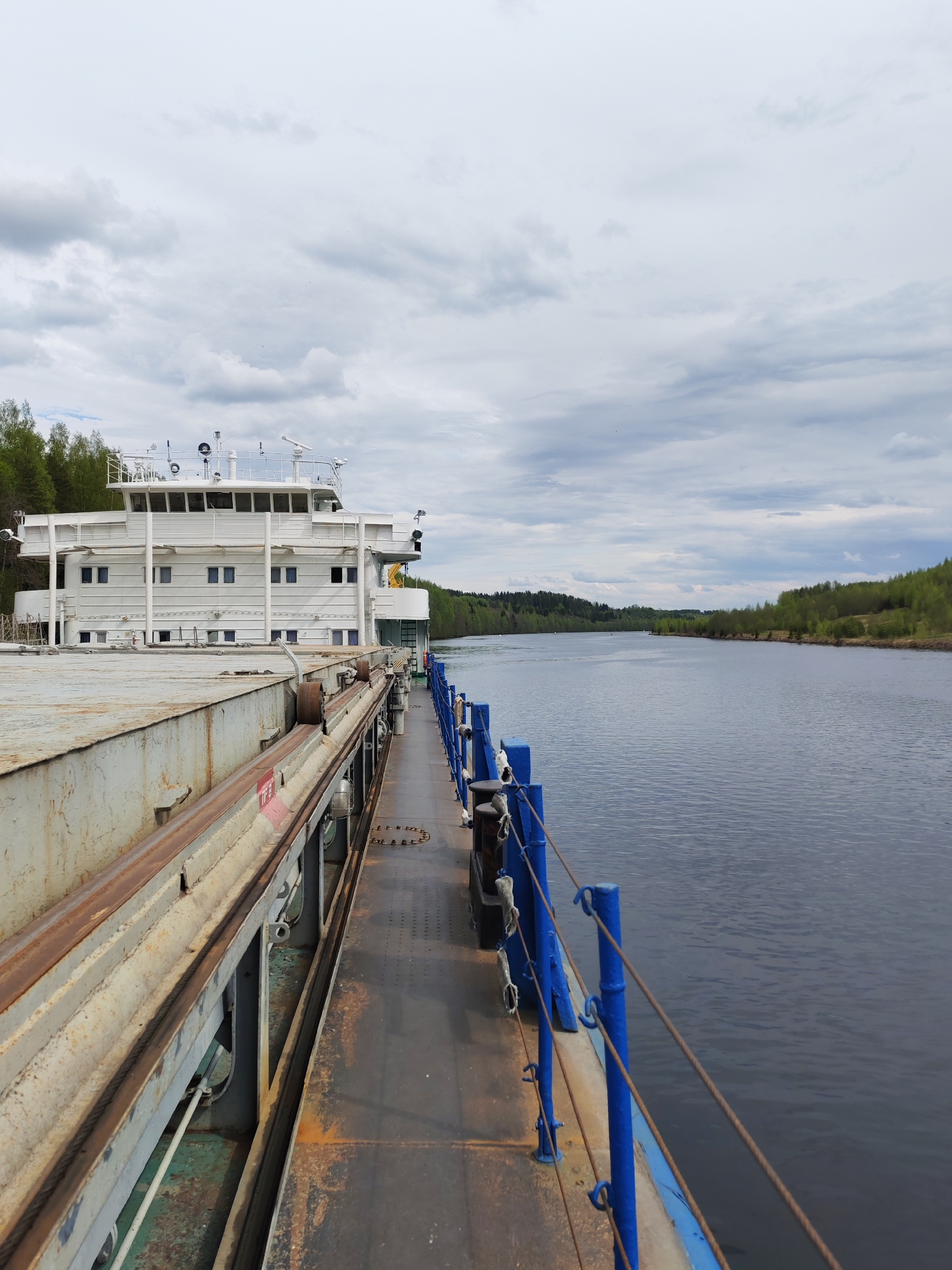 The image depicts a view from the deck of a river vessel, possibly a barge or a passenger ship, traveling along a waterway. The vessel's structure, including a white, multi-story cabin section with windows, is visible on the left side of the frame. A metal walkway with blue railings runs along the right side, adjacent to the water.
The body of water extends into the distance, bordered by forested areas on either side. The sky is overcast with grey clouds.
There is no verifiable indication of the location being Alekseevskoe, Russia, within the frame. No people are visible.