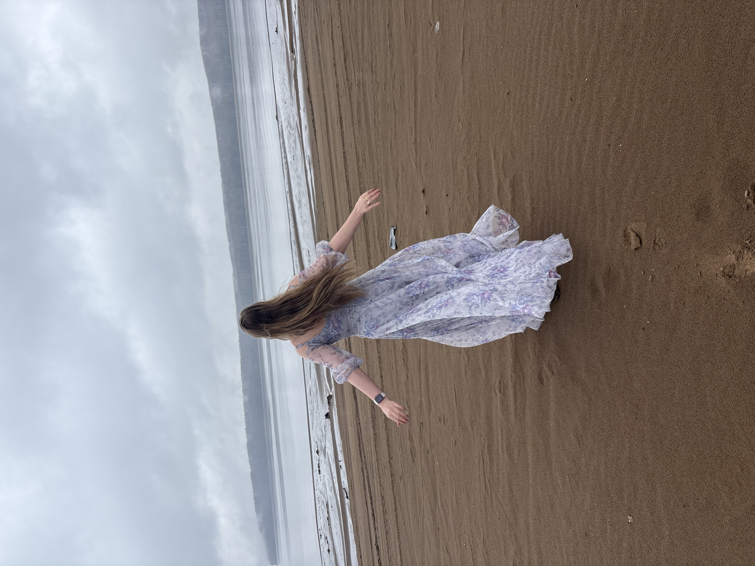 The original image is rotated 90 degrees clockwise, depicting a person lying on their back on a wide, sandy beach in Chaikovsky, Russia. The individual has long, light brown hair fanned out around their head and is wearing a flowing, light-colored dress with a subtle floral pattern, possibly white or pale purple. Their arms are outstretched to their sides, and a dark smartwatch is visible on their left wrist.
The setting is a broad, wet sandy or muddy shoreline next to a calm body of water, likely the Kama River or Votkinsk Reservoir. The water reflects the grey, overcast sky, indicating cloudy daytime weather. In the distance, a dark, indistinct landmass, possibly forested hills, stretches across the horizon beyond the water. The person's pose, with arms open, suggests a moment of relaxation, surrender, or artistic expression. No visible text is present in the image.
