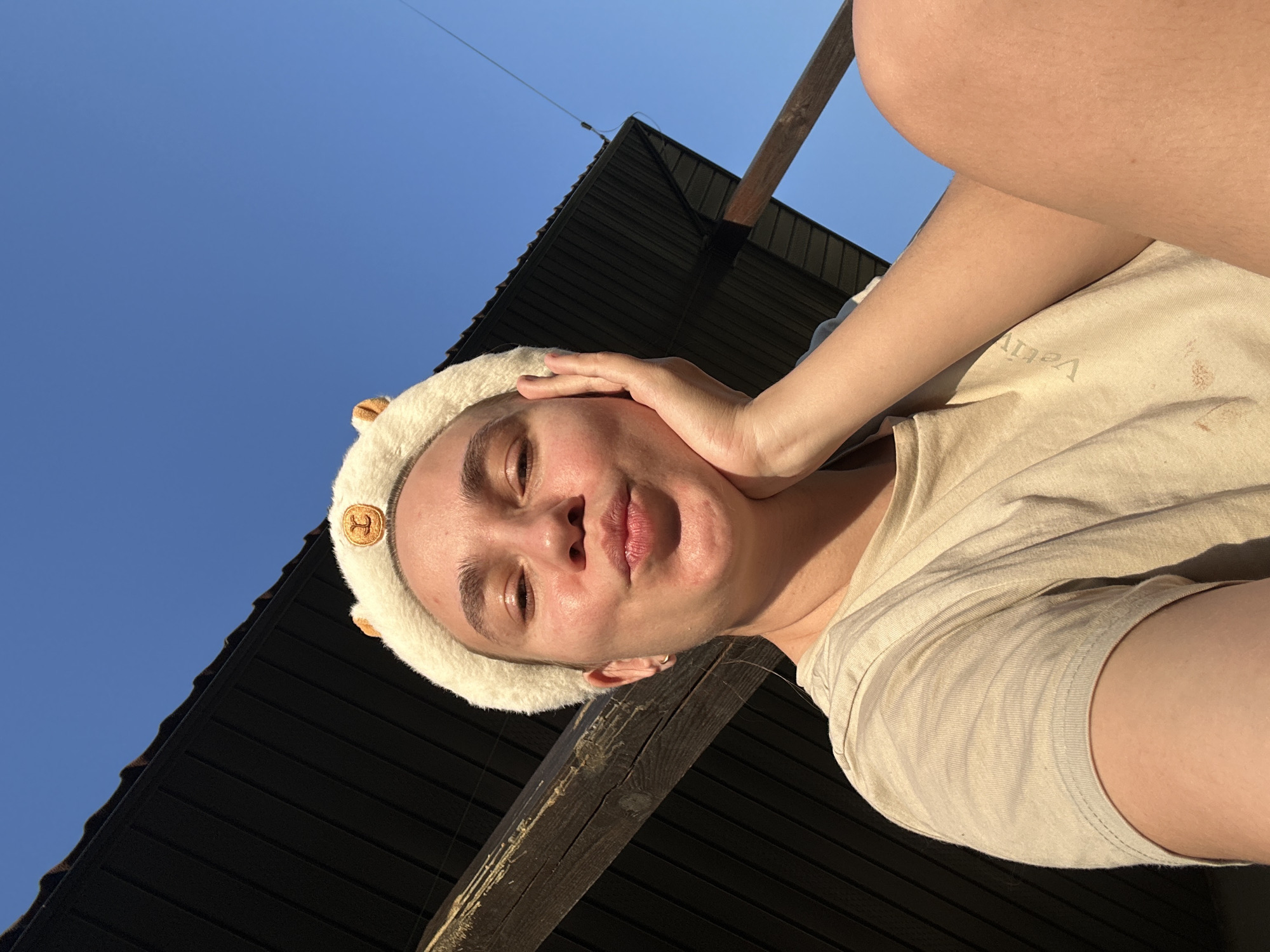 A person wearing a fluffy headband with circular decorations stands outside on a sunny day in what could be the city of Kharnavy, Russia. A building with dark siding and a wooden beam are behind the person. They have a light-colored shirt on and are holding their hand to their face. The sky is a clear blue.