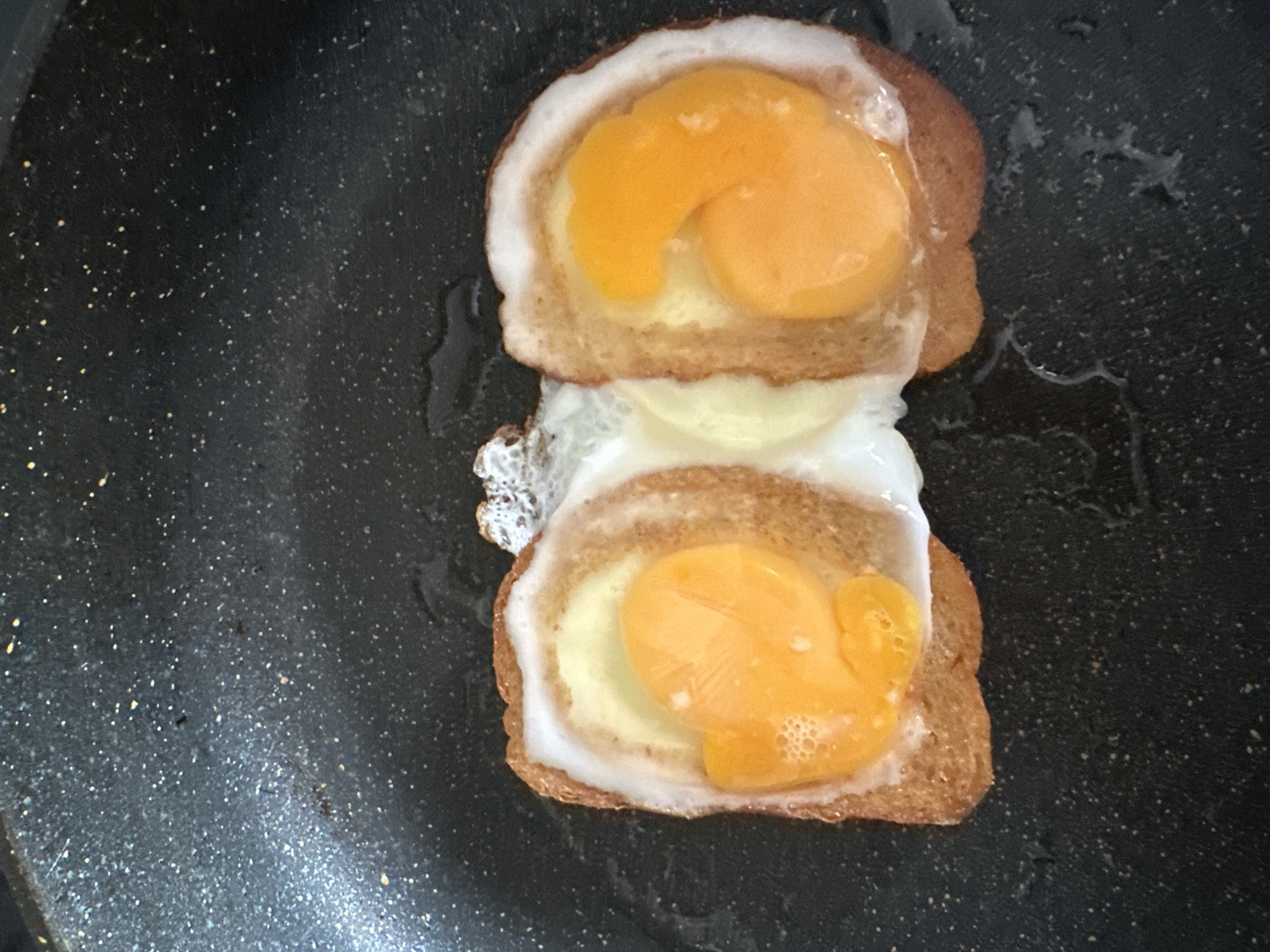 The image shows two fried eggs in toast rings on a black non-stick frying pan. The eggs have bright yellow yolks and white egg whites that have spread out. The toast rings are golden brown. The pan has a speckled surface and appears to have some oil or grease on it. There is no indication of Podolsk, Russia in the image, and no people or interactions are visible.