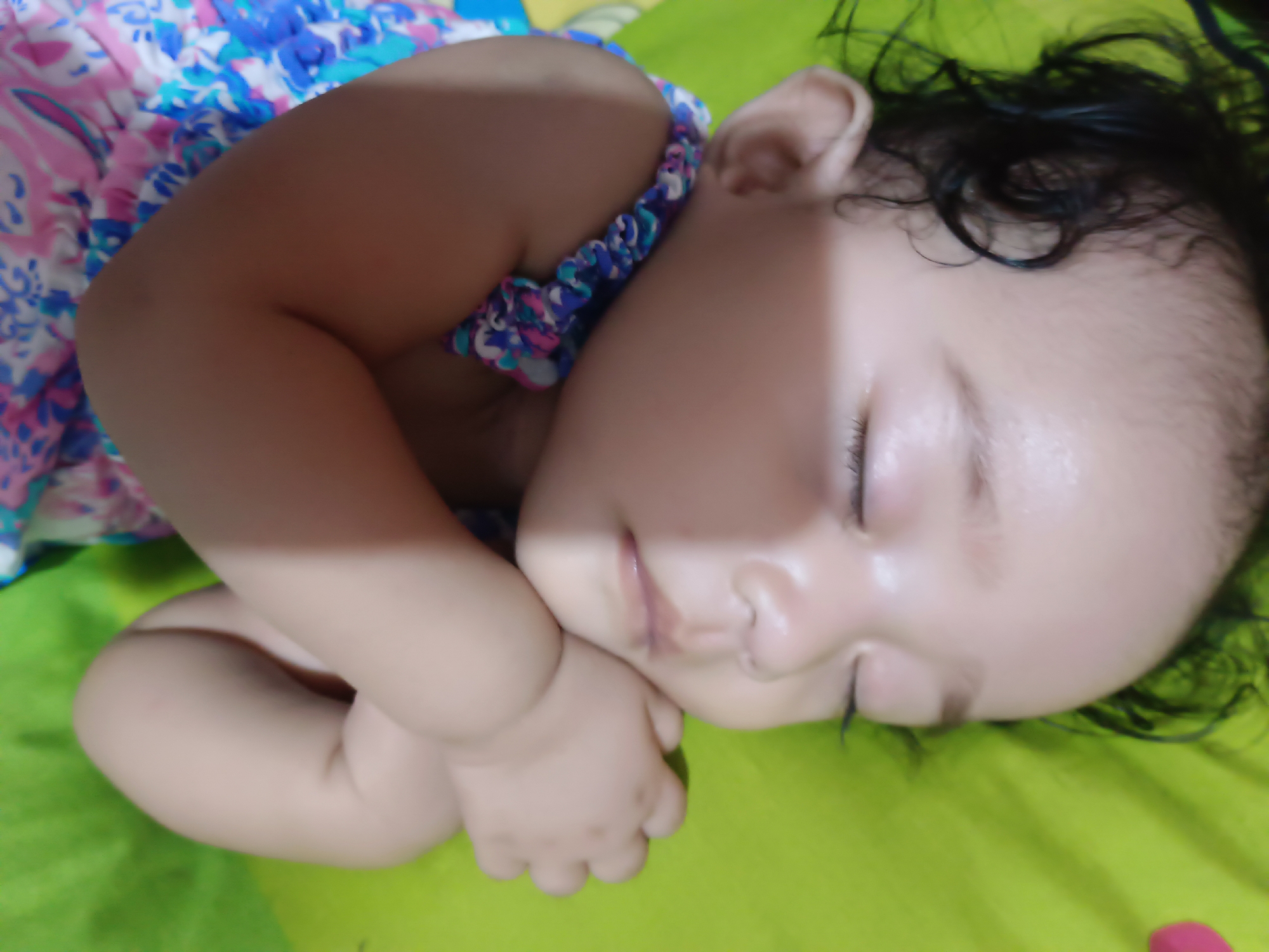 The image shows a close-up view of a baby sleeping on a green surface. The baby is lying on its side with its arms crossed in front of its chest. The baby is wearing a patterned dress with blue, pink, and white colors. The baby's head is turned to the side, and its eyes are closed. Its hair is dark and curly. A shadow is cast across the baby's face. The green surface appears to be a fabric, possibly a blanket or bedsheet. The image is taken from a high angle, looking down at the baby. There are no visible signs of the location being in the city of Kawua, Indonesia.