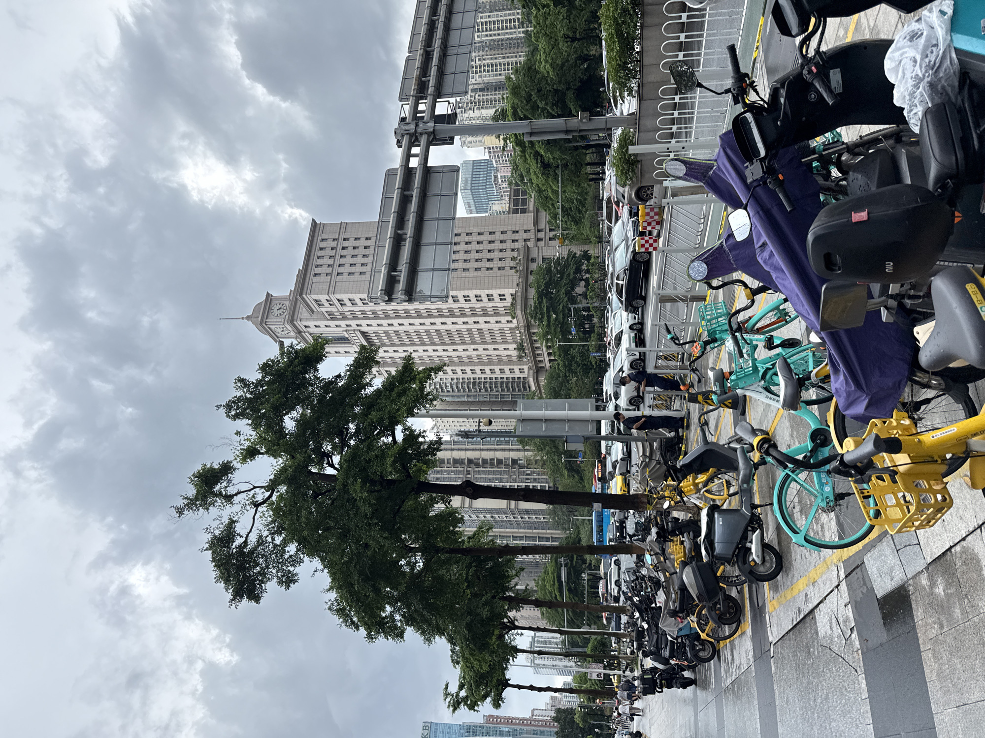 An overcast day in Tianhe District, China, features a street scene with parked motorcycles and bicycles lining the sidewalk. In the background, a prominent light-colored building with a clock tower rises above other high-rise structures. Traffic, including cars and a blue bus, moves along the road. Several trees with lush green foliage are planted along the sidewalk. Two men in dark uniforms stand near a white car, possibly security personnel or officials. The ground appears wet, suggesting recent rain.