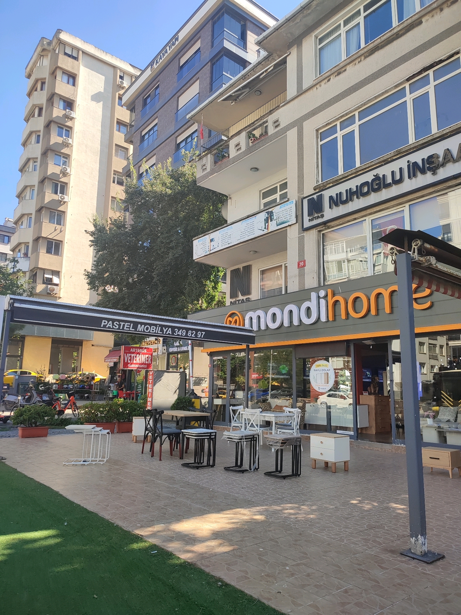 This image captures a street scene in Kadıköy, Turkey, under a clear blue sky. In the foreground, a patch of artificial green turf borders a tiled pedestrian area where several pieces of outdoor furniture are visible, including white chairs and tables, and a black metal chair with a matching table. A light-colored pole stands on the right side of the frame.
The midground features the storefronts of several businesses. A prominent orange and white sign reads "mondihome," indicating a furniture store. Adjacent to it, another awning displays "PASTEL MOBİLYA 349 82 97," suggesting a furniture shop as well. Further to the left, a red awning with white lettering announces "VETERINER," indicating a veterinary clinic. There are also various potted plants and some parked motorcycles and bicycles on the left side of the street.
The background is dominated by several multi-story buildings. A tall, beige apartment building stands on the left, with numerous windows and air conditioning units. To its right, a more modern building with gray and beige facades features dark-paneled windows. A sign on this building reads "NUHOĞLU İNŞAA." The architecture suggests a mix of residential and commercial spaces typical of an urban environment in Turkey. The overall impression is of a busy commercial street in a city.