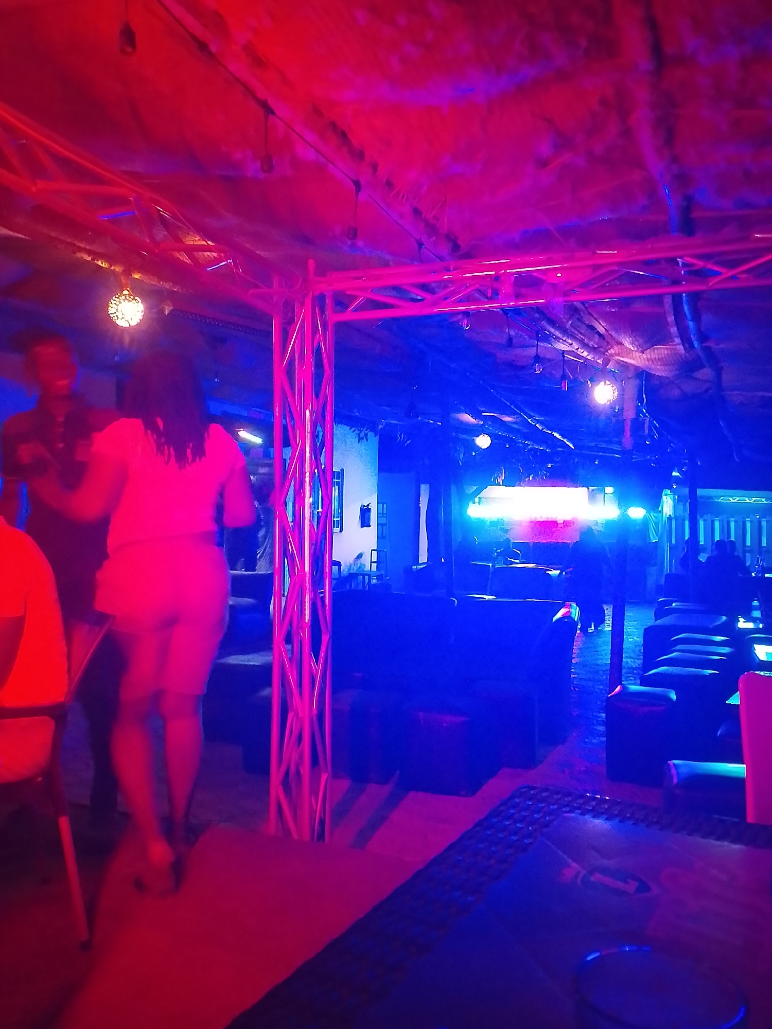 The image captures a vibrant, dimly lit interior scene, likely a club, bar, or lounge, characterized by strong red and blue neon lighting. The time of day is clearly night, as artificial lighting fully illuminates the space.
In the foreground and mid-ground, two figures are prominent on the left side of the frame. A man with dark skin, wearing a dark shirt, is partially visible. Standing with her back to the camera, a woman with lighter skin wears a light-colored top and shorts, her hair appearing dark and curly. They seem to be standing or dancing together. To the far left, part of a red chair is visible.
The setting features a distinctive structural element: a metallic truss system, glowing pinkish-red from the ambient lighting, stretches across the ceiling and descends vertically on the left side, possibly supporting lights or other decor. Several round, ornate light fixtures hang from the ceiling, casting soft glows. The ceiling itself appears to be covered, possibly with fabric or netting over a more basic structure.
Throughout the rest of the venue, dark-colored, plush-looking couches or cushioned benches are arranged, suggesting comfortable seating areas for patrons. In the background, other indistinct figures are visible, some appearing to be seated. A bright, rectangular light source, possibly a screen or stage lighting, is prominent in the distant background. Faintly visible on a white wall in the background is what appears to be a fire extinguisher. The floor is dark, with a textured mat visible in the lower right corner.
The overall atmosphere is lively and social, consistent with an entertainment venue. There is no clearly visible or legible text in the image.