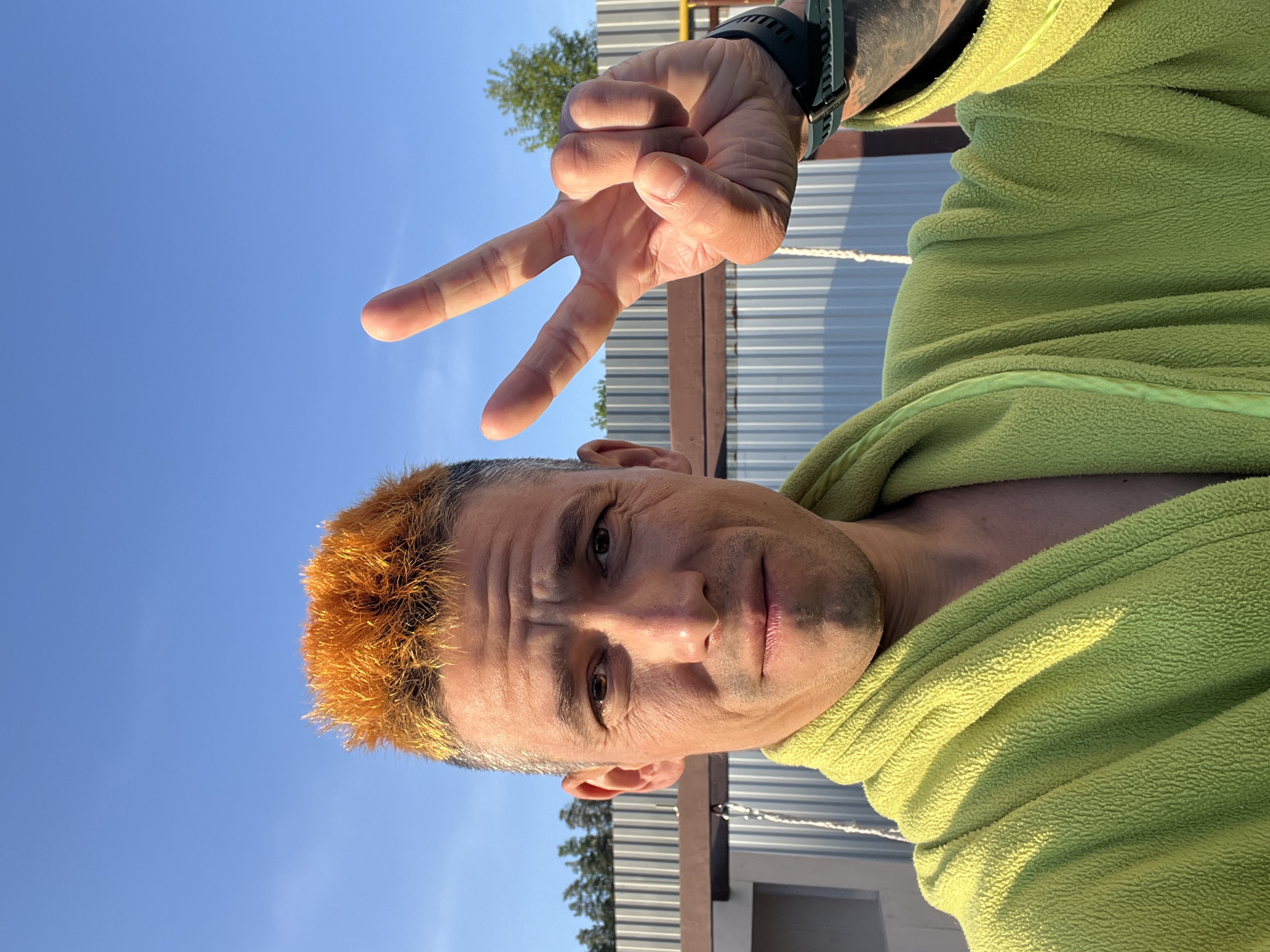 A man with orange hair stands outside in front of a metal structure, possibly a building or fence, with trees visible in the background. He is wearing a lime green fleece top and a dark-colored wristwatch on his left wrist. He is making a "peace" or "victory" sign with his hand. The sky is blue. The image appears to have been taken in an outdoor location, possibly in the city of Chaikovsky, Russia.