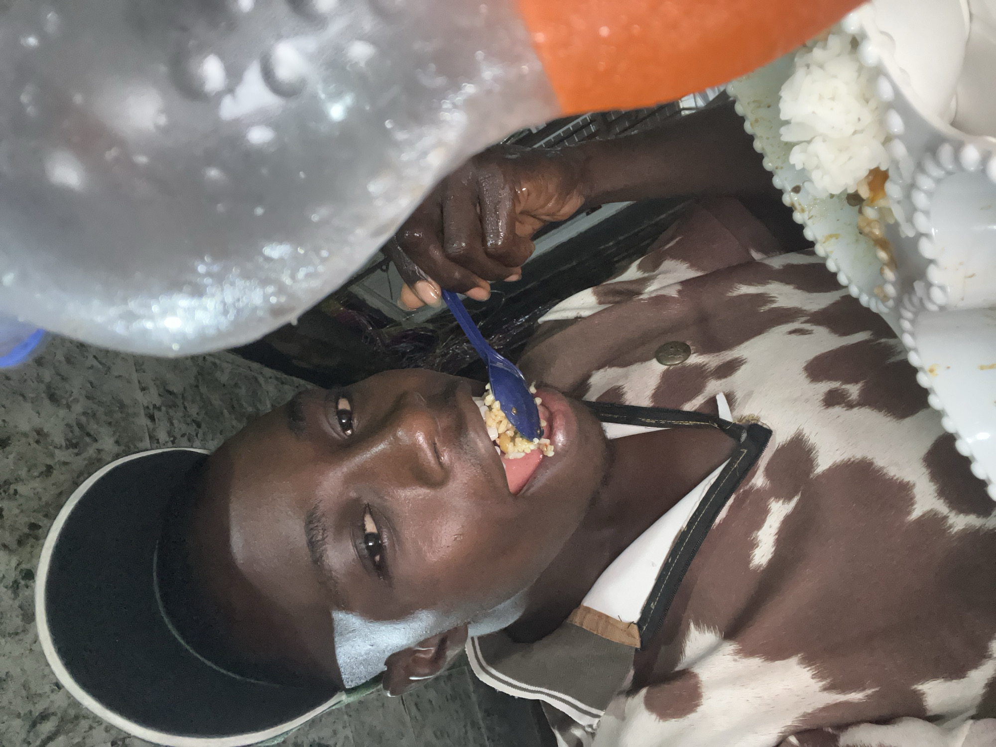 A young Black male is captured in a close-up, self-taken photograph, with his face dominating the foreground. He is wearing a dark baseball cap tilted backwards, revealing a portion of his forehead and eyes. His gaze is directed towards the camera, with his mouth open as he brings a spoonful of food to his lips. The food appears to be rice with some sauce or other ingredients. He is also wearing a shirt with a cow-print pattern. To the right of the frame, a translucent plastic bottle, partially filled with an orange liquid, is visible, suggesting it is a beverage. The background is dimly lit and appears to be an indoor setting, possibly a home or a small eatery. The overall impression is one of a casual moment, likely in Nkpor, Nigeria, where the individual is enjoying a meal.