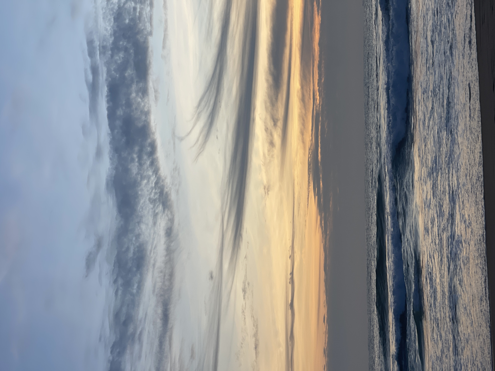 A wide-angle shot captures a tranquil ocean scene at sunset on Karon Beach, Thailand. The sky transitions from a soft blue at the top to hues of orange and yellow near the horizon, with wispy, streaky clouds adding texture. The sea is calm, with gentle waves lapping onto the sandy shore. The foreground shows the dark sand of the beach, meeting the water's edge. No people or objects are visible in the frame, emphasizing the serene natural beauty of the location.