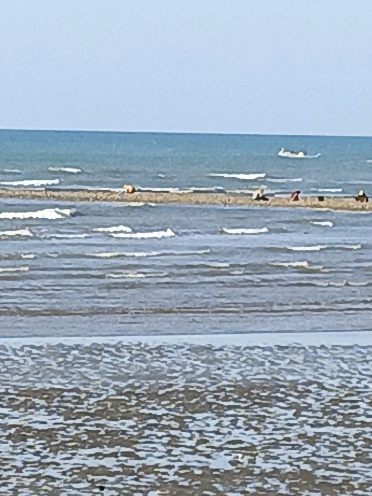 The image depicts a beach scene with a calm sea under a clear sky. In the foreground, a wet sand area with small pools of water indicates the receding tide. Further out, shallow water with gentle waves breaks over a sandbar. On this sandbar, several people are visible. One person appears to be wearing an orange top, while others are in darker clothing, some appearing to be sitting or lying down. In the distance, on the water, a white inflatable object with people on it is floating. The overall impression is a serene beach environment with people enjoying leisure activities. This scene is consistent with the beaches found in Bulukumba, Indonesia, known for its coastal beauty and leisure spots.