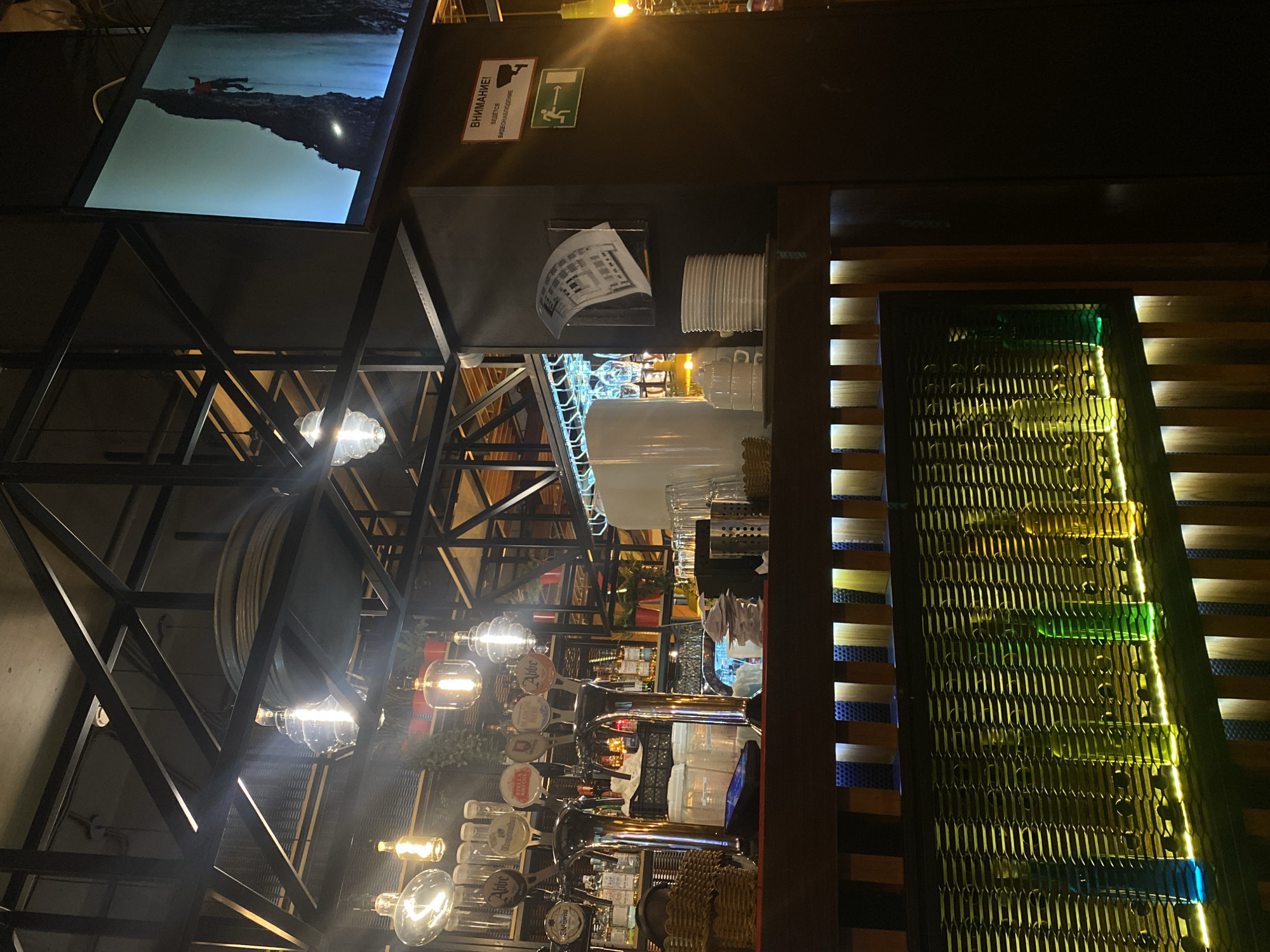 The image depicts the interior of a bar in Kazan, Russia. The scene focuses on the bar area, which features multiple beer taps with brand name emblems. Above the taps are stacked glasses, pitchers, and other bar equipment. Exposed light bulbs hang from the ceiling. Above the bar is a metal frame structure and a television screen displaying a dark image. Signage with Russian text and pictograms is visible. To the right, a decorative panel with colored accents adds to the ambiance.
