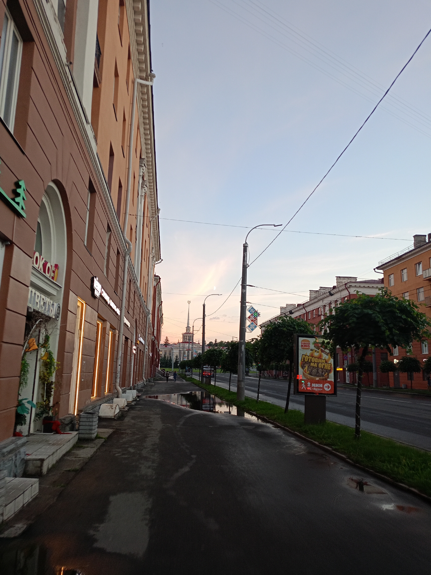 The image depicts a street scene in a city, likely in the evening or early morning given the soft lighting and the sky's hue. The perspective is from a sidewalk looking down a street lined with buildings on the left and a road with some trees and more buildings on the right.
On the left, a multi-story building with an orange-brown facade dominates the foreground. Its lower level features storefronts with illuminated signs, one of which reads "ОКОН" in large letters, suggesting a window-related business. Further down the sidewalk, there are more illuminated storefronts. The sidewalk itself is wet and reflects the lights, indicating recent rain or puddles.
On the right side of the road, there are several trees and more buildings. A prominent billboard advertising Burger King with the slogan "ГОТОВИМ НА ОГНЕ" (We cook over fire) is visible. Across the road, there are residential buildings with multiple windows. In the distance, a tall spire or tower is visible against the sky, possibly a landmark.
The sky is a gradient of pale blue and soft pink, suggesting a twilight or dawn setting. Several power lines crisscross the sky. Two figures are walking on the sidewalk in the distance. The overall impression is a quiet urban street scene after a period of rain.
