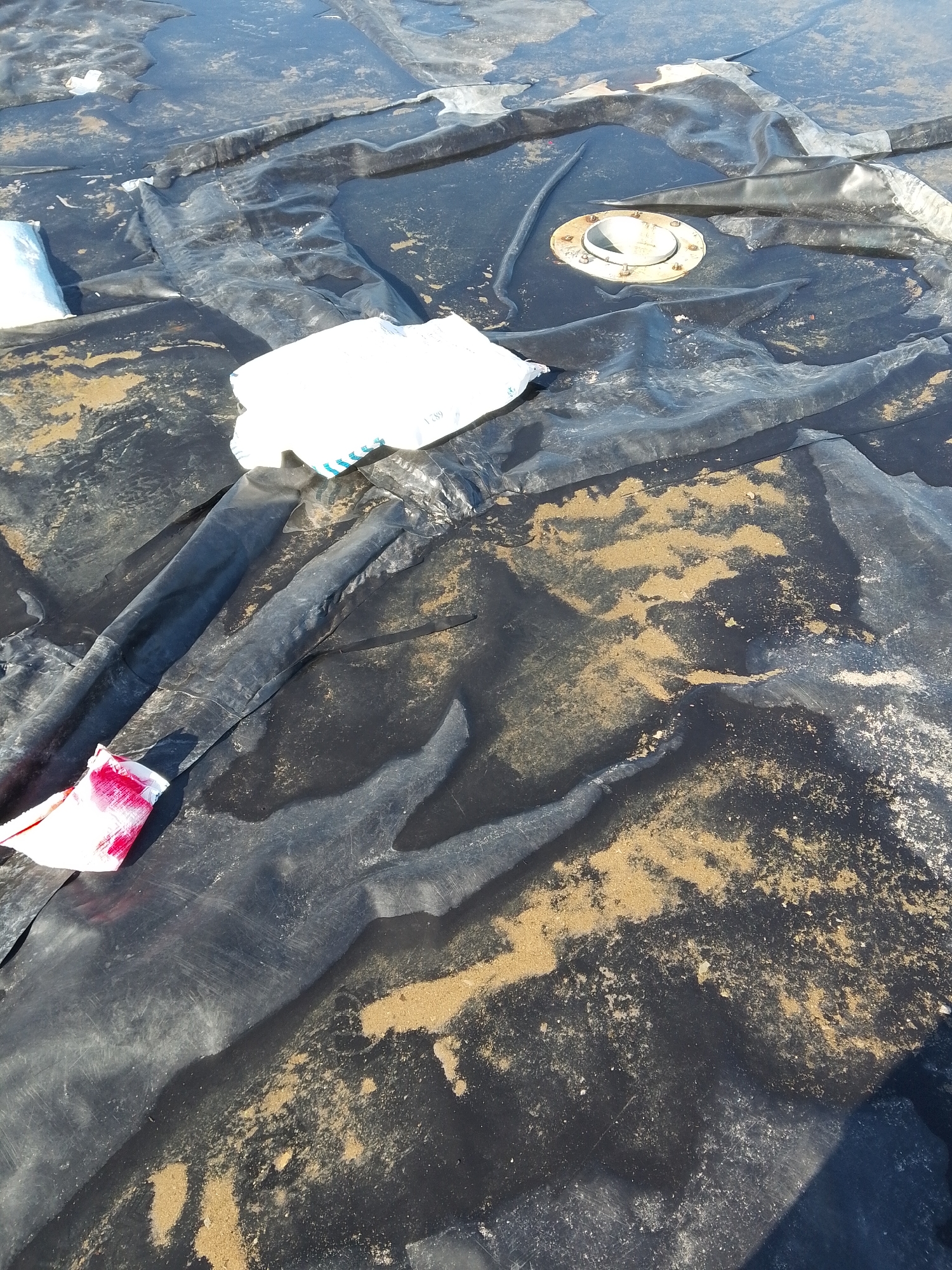 The scene depicts a section of a black, possibly plastic or synthetic, membrane or tarp surface. The surface is uneven, with folds, wrinkles, and pooled water in places. There are patches of sand or dirt scattered across the surface. Two white bags are on the tarp. One white rag with red staining is also present. In the top center, there is a metallic flange or fitting with visible bolts, likely part of a tank or pipe. The location is in the city of Bukit Ketok, Indonesia.