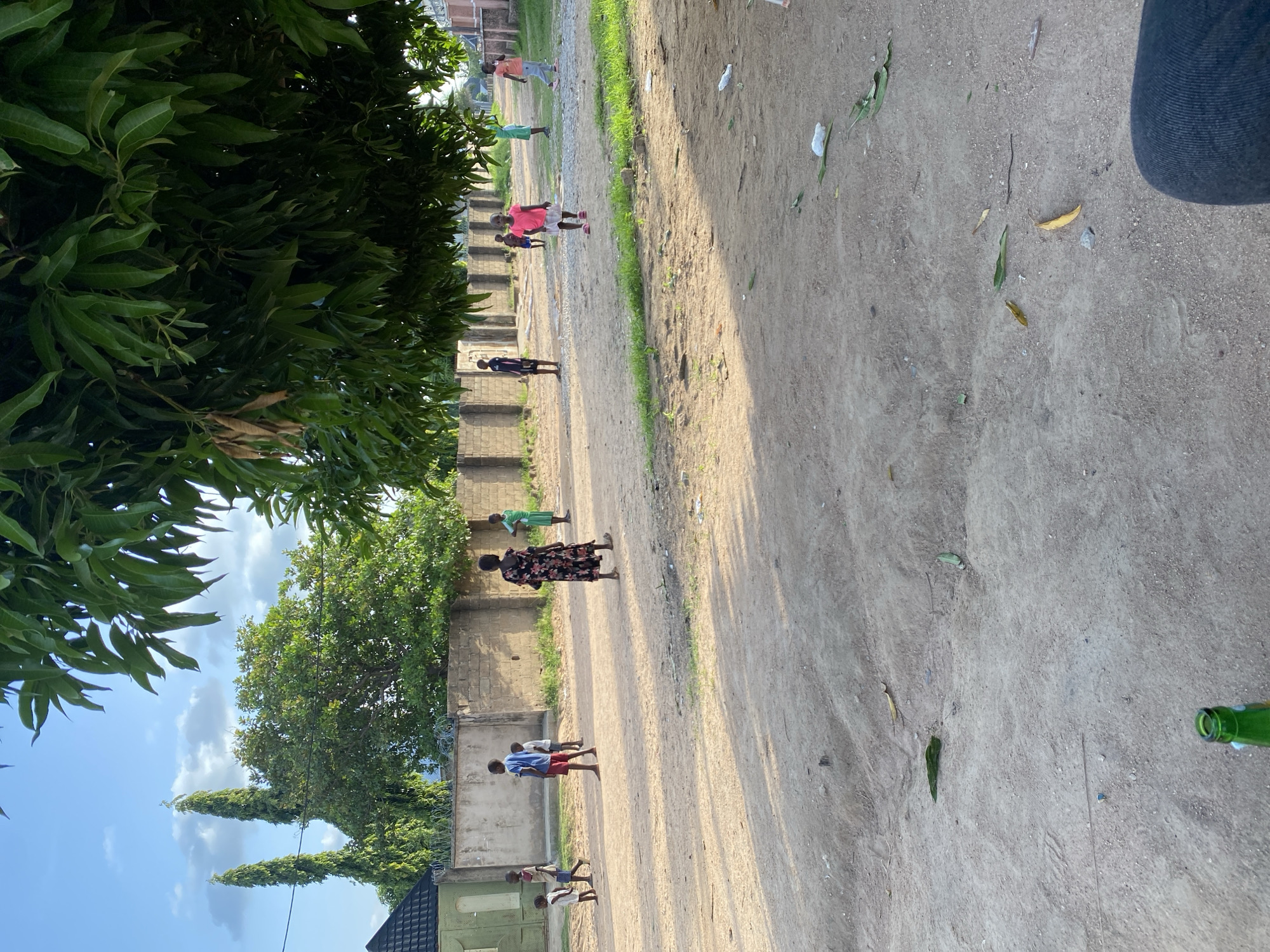 A group of children are playing in a dirt lot in Jalingo, Nigeria. The lot is bordered by a low brick wall and fences. Trees with large green leaves overhang the scene from the right. The sky is blue with scattered clouds. The children are of various ages and are wearing casual clothing. One child in the foreground is wearing a bright pink shirt and a white skirt. Another child in the background is wearing a dark floral dress. A few other children are visible in the distance, some wearing blue shirts and red shorts, and others in green shirts. The ground is dry and dusty, with patches of sparse grass. There is some litter scattered on the ground, including white plastic bags and leaves. In the foreground, the top of a green bottle is visible.