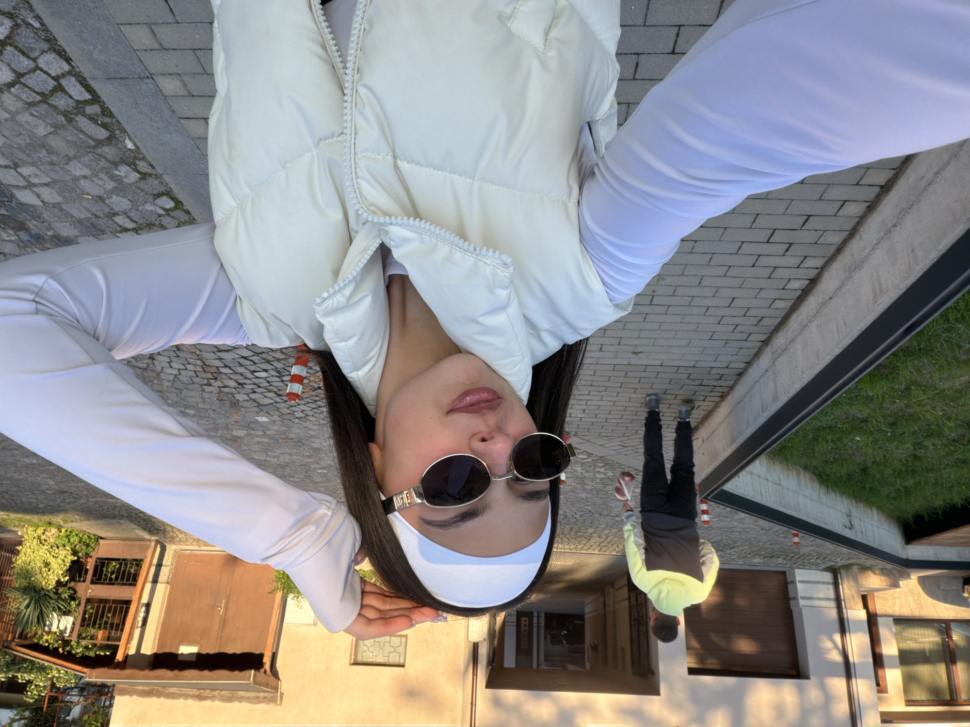 A young woman, possibly in her early twenties, is posing for a selfie. She is wearing a white padded vest over a long-sleeved white top, a white headband, and sunglasses. A second person, a young man, is walking behind her, holding what appears to be a red bag or bottle. The background features a residential area with buildings, stone pathways, and greenery. Based on the architectural style and pathway material, the location may be in Gabrovo, Bulgaria.