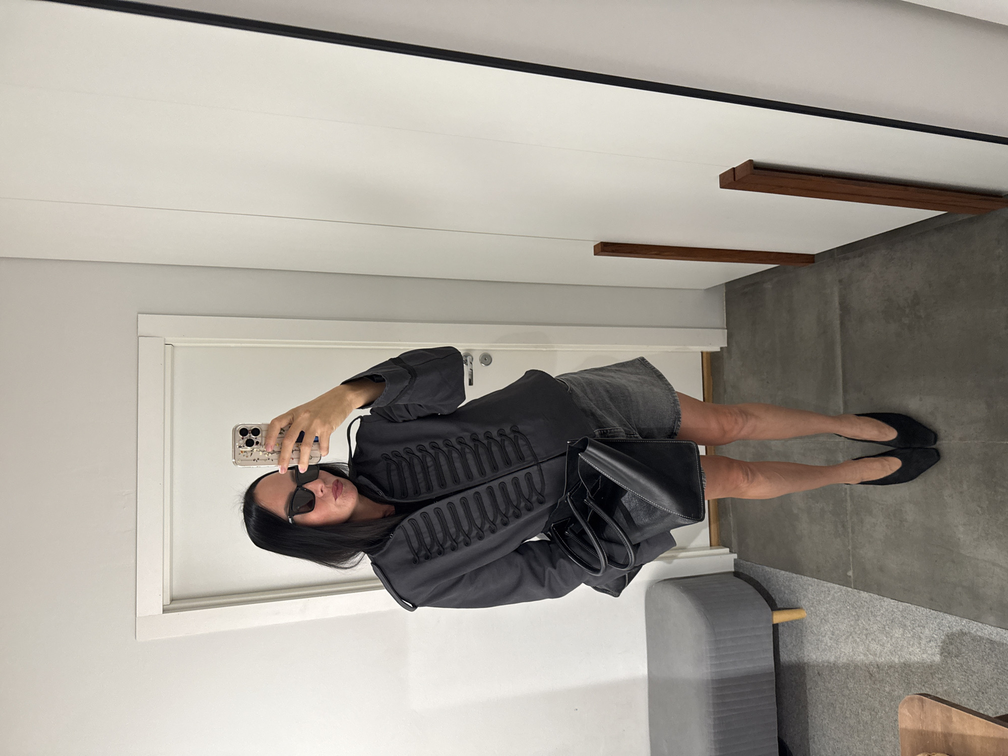 A woman stands in a brightly lit room, taking a mirror selfie with her phone. She is wearing a dark gray, military-style jacket with ornate black trim, a dark t-shirt, and a gray denim skirt. She also has on black pointed-toe heels and dark sunglasses. A black tote bag rests on her left arm. The room appears to be a dressing room or a fitting area, with a large white wardrobe or door to her right, a white door with a silver handle to her left, and a gray upholstered bench with wooden legs in the background. The floor is a polished concrete or tile. There is no indication that this scene is located in Chaikovsky, Russia.