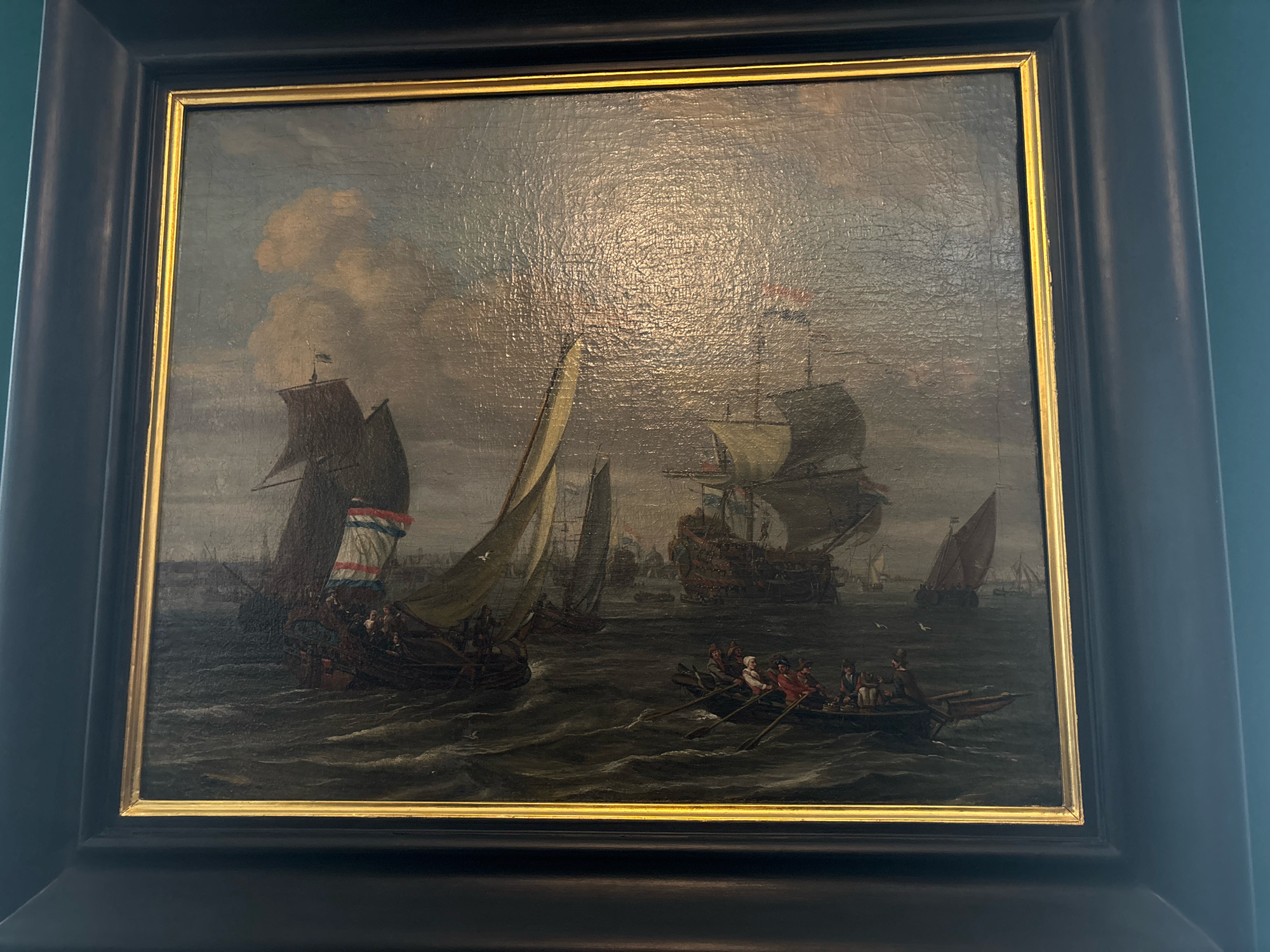 This is a medium shot, eye-level photograph of a framed oil painting depicting a historical maritime scene. The painting is enclosed in a dark, wide, wooden frame with a narrower gold frame directly around the artwork. The artwork itself is a detailed depiction of several sailing vessels on a choppy, dark gray sea under a cloudy, atmospheric sky.
The main subject is a fleet of ships, with a large galleon-style ship prominently featured in the mid-ground, to the right of center. This ship has multiple sails and appears to be a man-of-war. To the left of the galleon, another large sailing vessel with tan sails is visible, carrying people and displaying a flag with red, white, and blue horizontal stripes. In the foreground, a smaller rowboat with several figures dressed in historical attire is depicted, actively rowing through the water. The figures appear to be men, some wearing hats. Smaller sailing boats and a distant coastline with buildings are discernible in the background, suggesting a port or busy waterway. The painting style is realistic, with attention to the texture of the water and the rigging of the ships. The lighting suggests an overcast day.
The painting is displayed on a dark green wall, likely in a museum or gallery setting. Based on the architectural style and the presence of numerous Dutch Golden Age paintings depicting maritime scenes, this painting is likely located in the Hermitage Museum in Saint Petersburg, Russia.