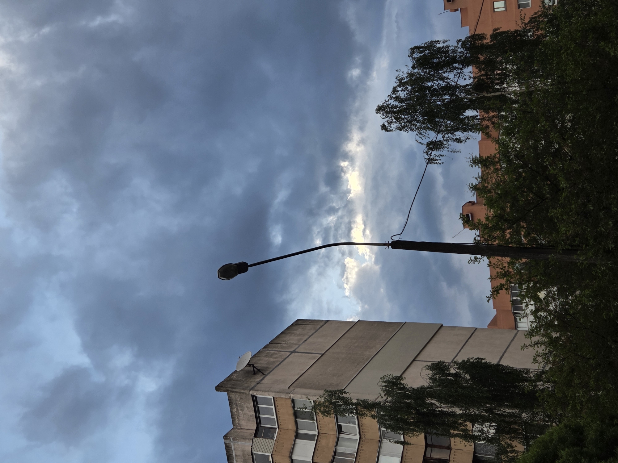 The image shows an upward view of a street scene in an urban environment. The sky is predominantly filled with dark, stormy-looking clouds, with a strip of light visible between them. A street lamp stands prominently, its pole extending upwards and the lamp head oriented to the left. Below, a multi-story residential building is visible, featuring multiple rows of windows and a satellite dish on the roof. The building's exterior is characterized by its worn, concrete facade. In the background, the upper part of another building, painted in a muted orange tone, is partly obscured by dense tree foliage. The foliage creates a natural screen that softens the urban environment. The overall impression is one of an overcast day in a densely populated area. The location is in the city of Unknown, Unknown.