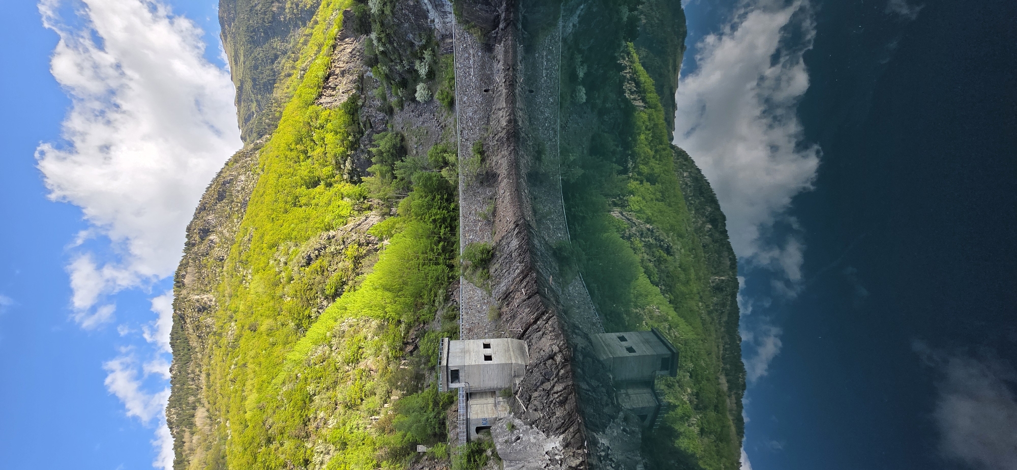 The image depicts a landscape scene in Benasque, Spain, featuring a mountain partially covered with lush green vegetation under a blue sky with scattered white clouds. The lower portion of the image shows a reflection of the mountain and sky in the water, indicating a lake or reservoir. A concrete structure, possibly a water intake or control building, is situated on the rocky edge between the mountain and the water. A narrow paved path runs along the side of the mountain. The reflection and orientation give the illusion of the scene being upside down.