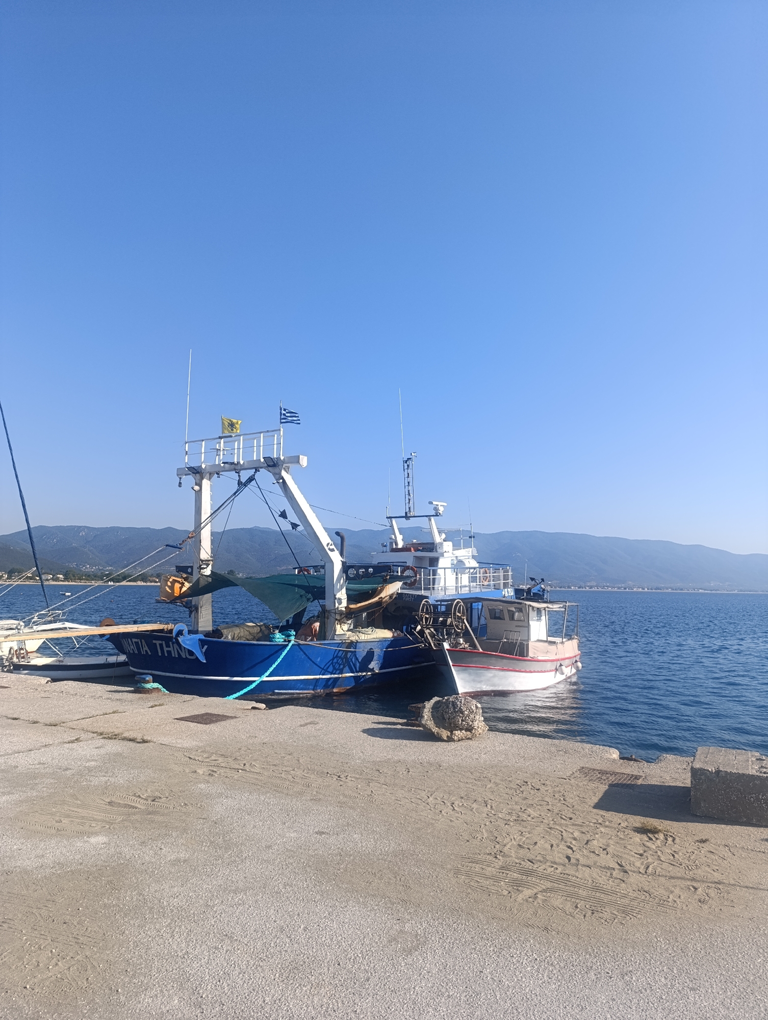 In the city of Stavros, Greece, a serene scene unfolds at a harbor. The clear blue sky stretches above, meeting the tranquil blue waters of the sea. A concrete pier, lightly sprinkled with sand and showing faint tire tracks, forms the foreground. Moored to the pier are two boats. The larger, a blue fishing vessel, prominently displays the name "MAFIA THINOY" in white lettering. It is equipped with various fishing gear, including a white crane-like structure and a net. Beside it, a smaller white and red boat is also docked. In the background, a lush green mountain range lines the horizon, dotted with scattered buildings, suggesting the presence of a coastal town. The overall impression is one of peaceful coexistence between human activity and the natural beauty of the Greek coast.