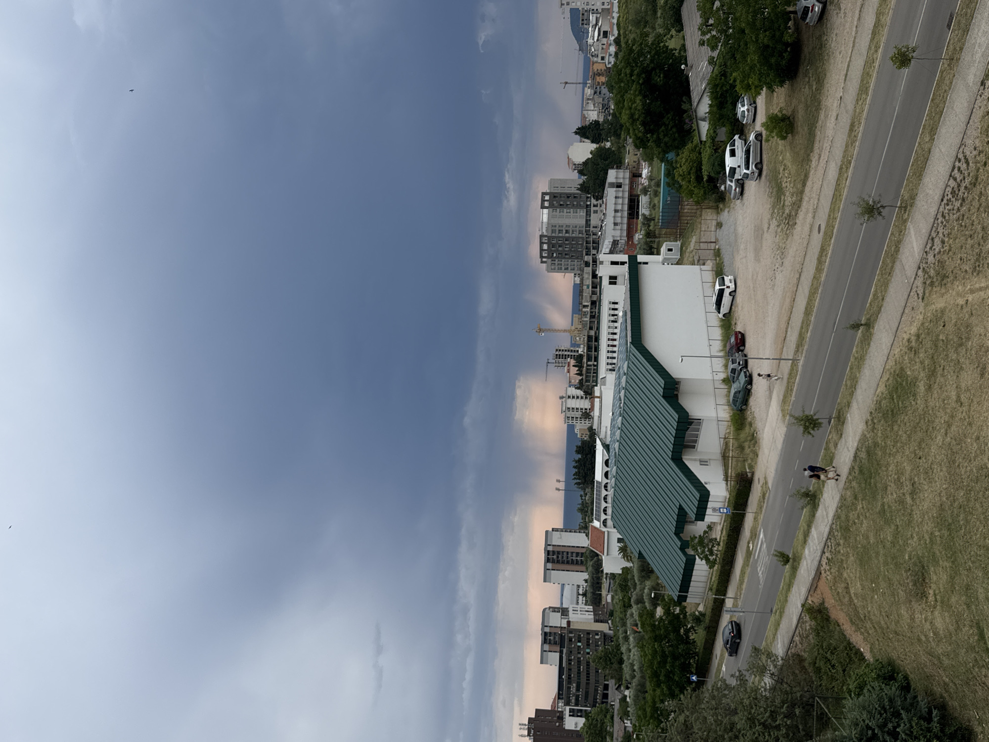 This image captures an overcast day in Bar, Montenegro, likely late afternoon given the soft, diffused light and hints of pinkish hues in the distant sky. The scene is a mix of urban development and natural elements.
In the foreground, a building with a distinctive green, sloped roof is prominent. Adjacent to it, a road with a single lane in each direction runs diagonally through the frame. Several cars are parked along the roadside and in a dirt lot. Two people are walking on the sidewalk next to the road, one appearing to be assisting the other. Further down the road, a black car drives away from the viewer.
The middle ground reveals a collection of apartment buildings and construction sites, with several cranes visible, indicating ongoing development. Trees, some with olive-like leaves, dot the landscape and line the streets.
The background is dominated by a vast expanse of sky, transitioning from a deep, moody blue on the left to lighter, cloudier conditions with a touch of orange and pink on the right, suggesting a potential sunset or the approach of a weather change. A small, dark speck in the sky might be a bird. The overall impression is one of a coastal city experiencing a period of growth under a dramatic sky.