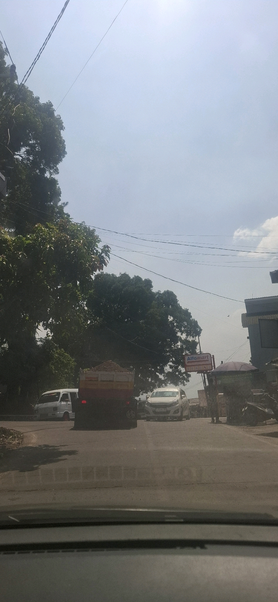 The image shows a view from inside a vehicle, looking out at a street scene in Cilaku, Indonesia. The sky is bright and mostly clear with some scattered clouds. Overhead, power lines crisscross the sky, casting shadows on the road. Lush green trees line the left side of the road, with some taller trees visible in the background.
On the road, several vehicles are present. A white van is parked on the left side. In the center of the image, a red and yellow truck is driving, carrying a load of dirt. To the right of the truck, a white car is also driving. Further to the right, a sign for "Alfamart" is visible, a common convenience store chain in Indonesia. Next to the Alfamart, a covered stall or structure is set up, with at least two individuals standing near it. A motorcycle is parked on the far right. The road surface appears to be asphalt. The overall impression is of a typical daytime street scene in a populated area.