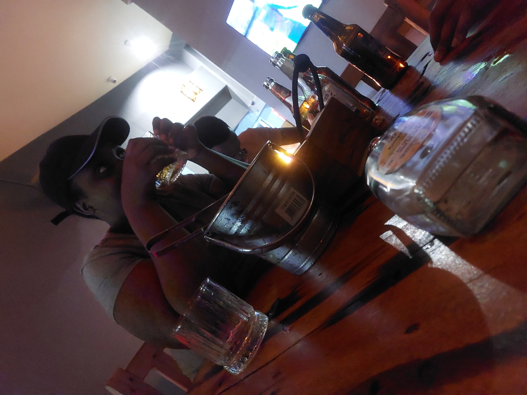 Here is a description of the image:
**Setting:** The image appears to be taken inside a bar or pub. The lighting is dim, with a warm reddish hue. There's a wooden table as the main surface. The location is Dar es-Salaam, Tanzania.
**People:** The primary subject is a person wearing a dark baseball cap. They seem to be in the act of eating something, possibly holding a piece of food in their hand. There's also another person partially visible in the background.
**Objects:** There are several bottles of beer on the table, along with a metal ice bucket containing more bottles. A glass with some liquid in it sits near the edge of the table. There is also a box of matches.
**Actions & Interactions:** The main action is the individual eating or about to eat. There is a social element suggested by the presence of multiple drinks and the other person.