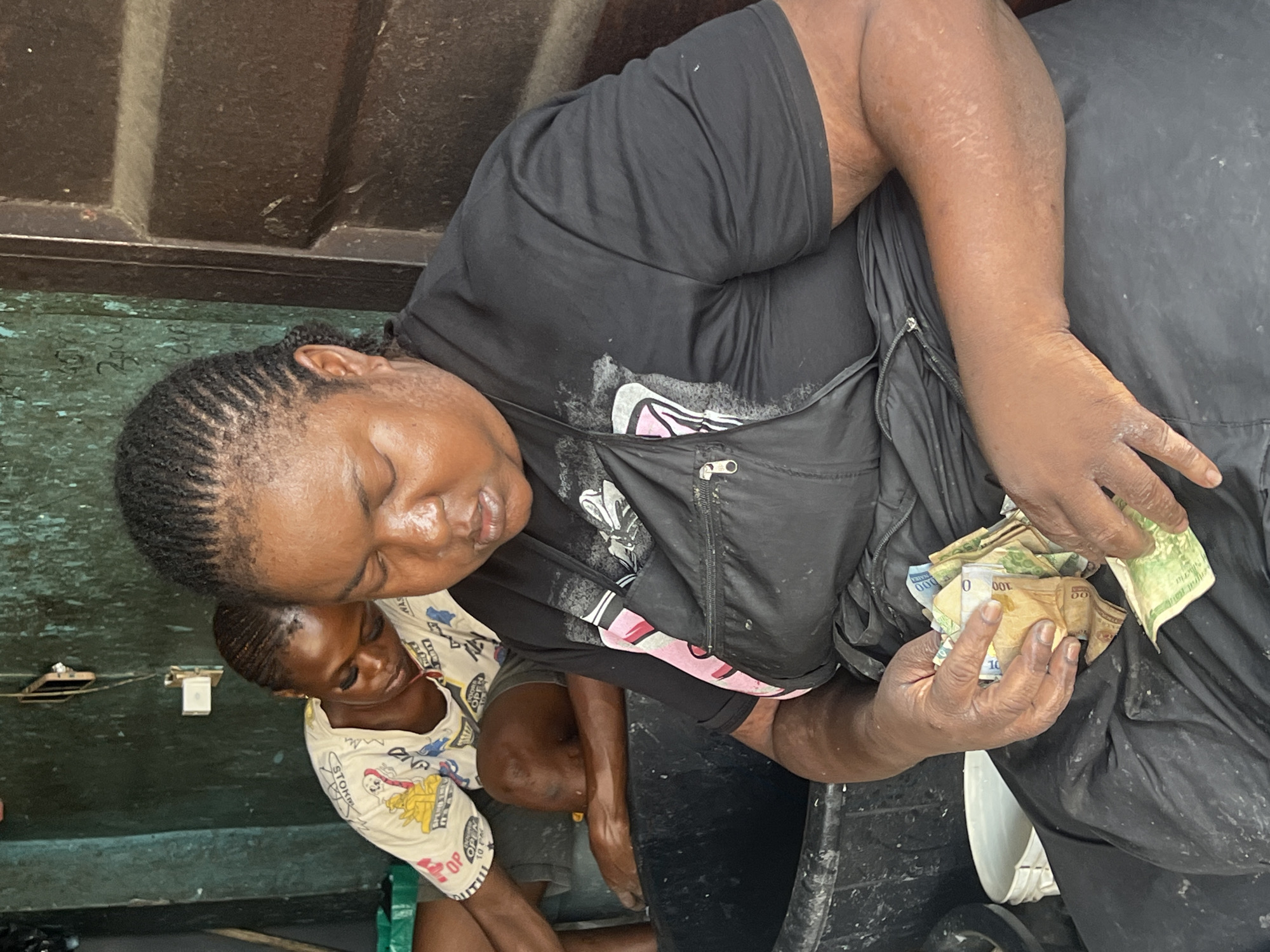 A medium shot captures a woman, presumably inside a building, counting a wad of Nigerian Naira notes. She wears a black shirt and vest. Beside her, a child in a white, cartoon-patterned shirt looks on. The setting is undefined, with a gray and brown ceiling above and a light green wall with electrical components behind the child.