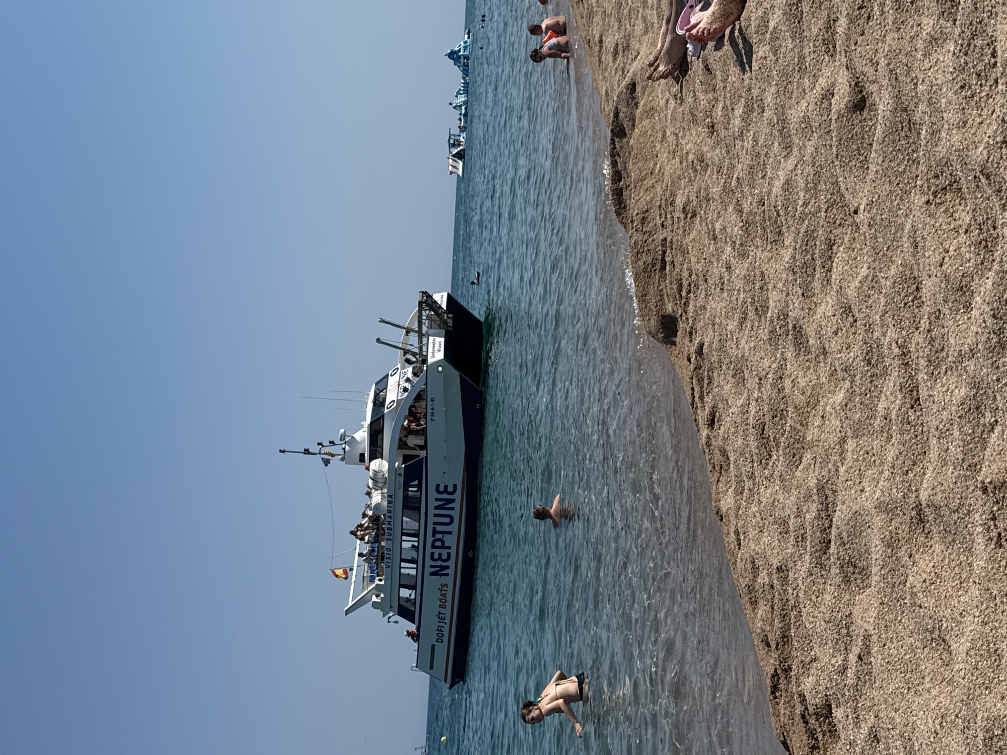 On a clear, sunny day at a beach in Blanes, Spain, a large tour boat named "NEPTUNE" with "DOFI JET BOATS" and "VISIO SUBMARINA" visible on its side is anchored in the shallow, blue water. Several people are seated on the boat's upper deck, and more are visible on the lower deck. In the water near the boat, a person is swimming. Closer to the shore, two young children are sitting at the edge of the water. Further out in the water, there are inflatable water attractions. The foreground is a sandy beach with footprints and some people's legs and feet visible. The sky is clear and blue.